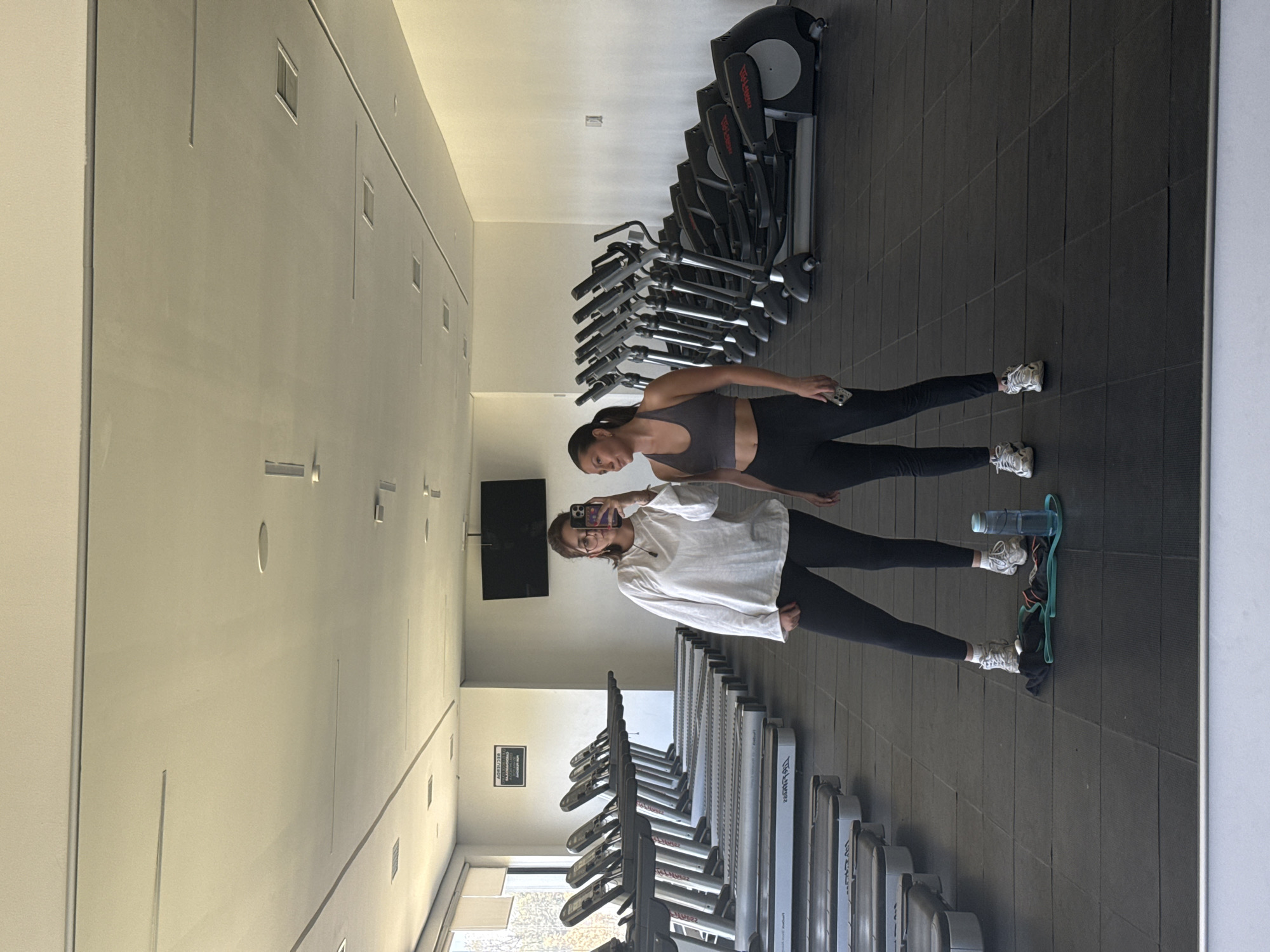 In a mirrored perspective at a gym in San Andrés Cholula, Mexico, two women are visible. One woman, wearing a sports bra and leggings, stands slightly behind the other. The second woman stands in front, wearing a white long-sleeve shirt over her workout clothes, holding a phone in her left hand to take a photo in the mirror. Behind them are rows of treadmills and elliptical machines. A water bottle and resistance band are on the floor. The floor is covered in dark grey squares. A television is mounted on the wall. The walls are light grey.