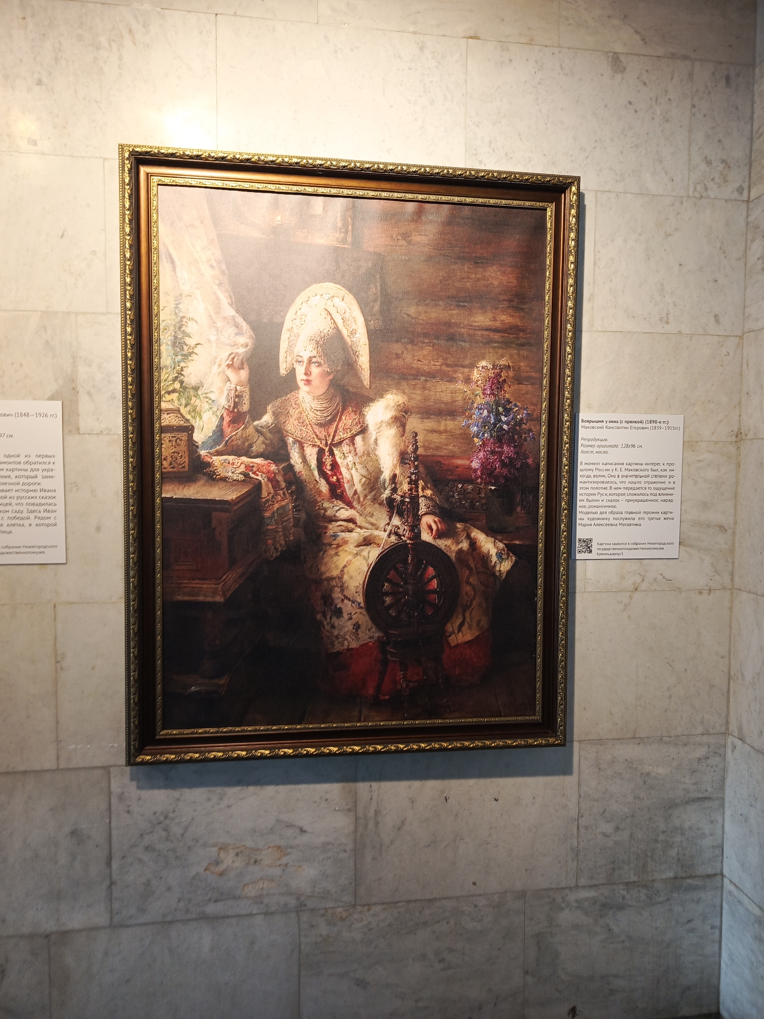 The image depicts a framed painting of a Russian woman, likely a noblewoman, seated and dressed in traditional clothing with a headdress. She is near a spinning wheel, with flowers and a chest visible. To the sides of the painting are informational plaques written in Russian about the painting and artist. The painting is mounted on a wall covered with marble tiles. The painting is “Boyar Woman at the Window” by Konstantin Makovsky. The painting is housed in the state art museum in the Kremlin in Nizhny Novgorod.