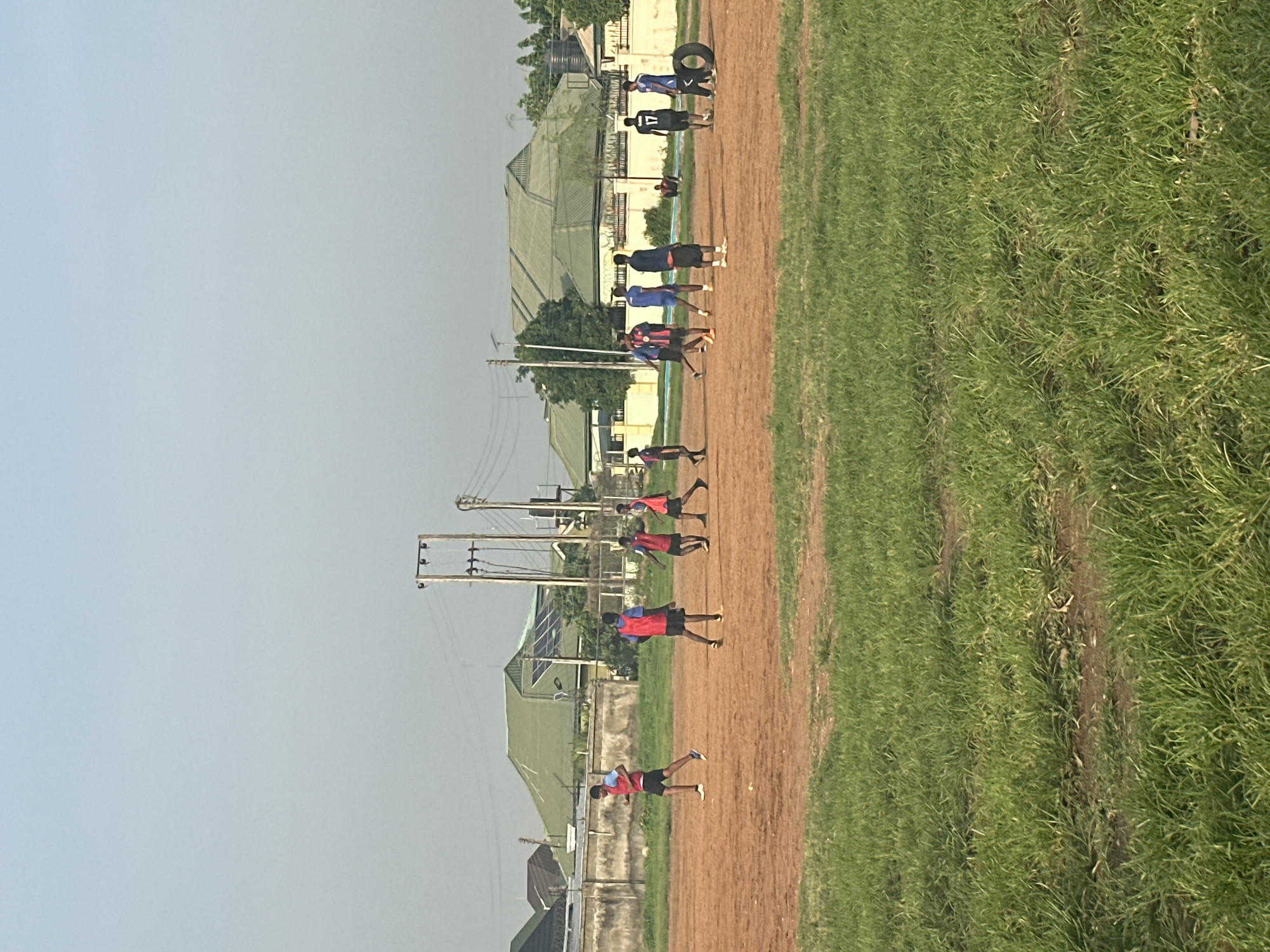 The image depicts a group of young men engaged in what appears to be a sports training session on a dirt track adjacent to a grassy field. They are dressed in athletic wear, with some wearing red vests over blue shirts, suggesting they are part of a team or group. The individuals are spread out along the track, some in mid-stride as if running or practicing drills, while others are standing. In the background, there are buildings with green roofs, trees, and utility poles, indicating an outdoor setting, likely a sports facility or school grounds. A tire is visible near one of the buildings. The sky is overcast and appears light blue, suggesting it is daytime, possibly late afternoon given the lighting. There is no visible text in the image. The overall scene conveys a sense of physical activity and training.