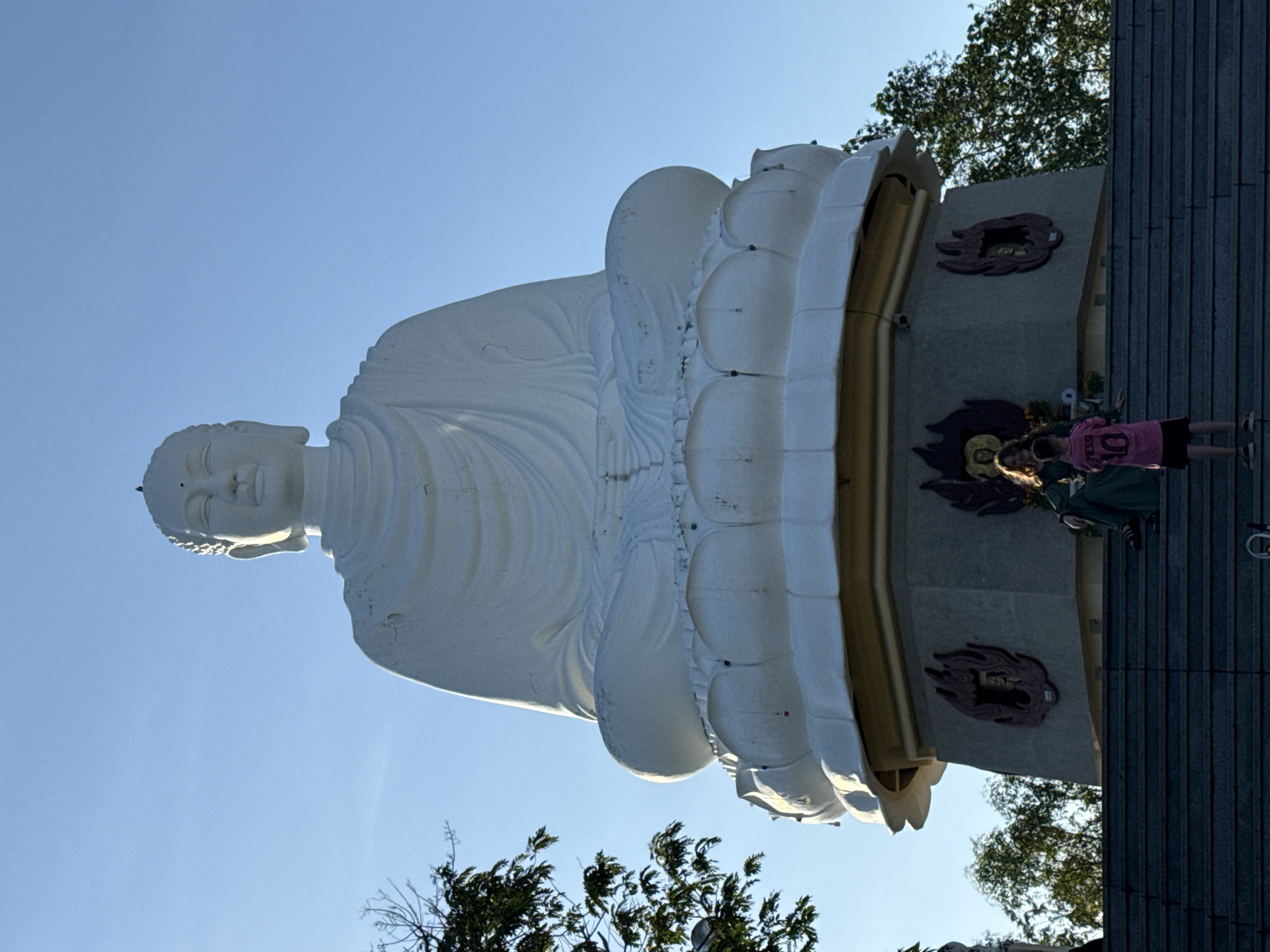 A low-angle shot depicts a large, white Buddha statue under a clear blue sky. The statue sits on a white, lotus-shaped pedestal trimmed with gold. At the base of the statue, two individuals are present, with one visible in a pink shirt and a second partially obstructed. The background also includes foliage and architectural elements. The location is suggested to be in Tay Nha Trang Ward, Vietnam.