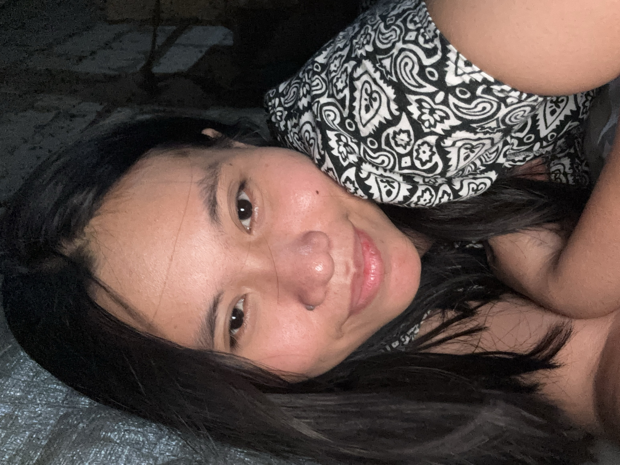 A young woman is lying down and taking a selfie. She has long, dark hair and is wearing a black and white patterned top. Her head is tilted to the side, and she is resting her cheek on her arm. The background is dark and appears to be an indoor setting. There are no specific landmarks visible to determine the location within East Jakarta, Indonesia.