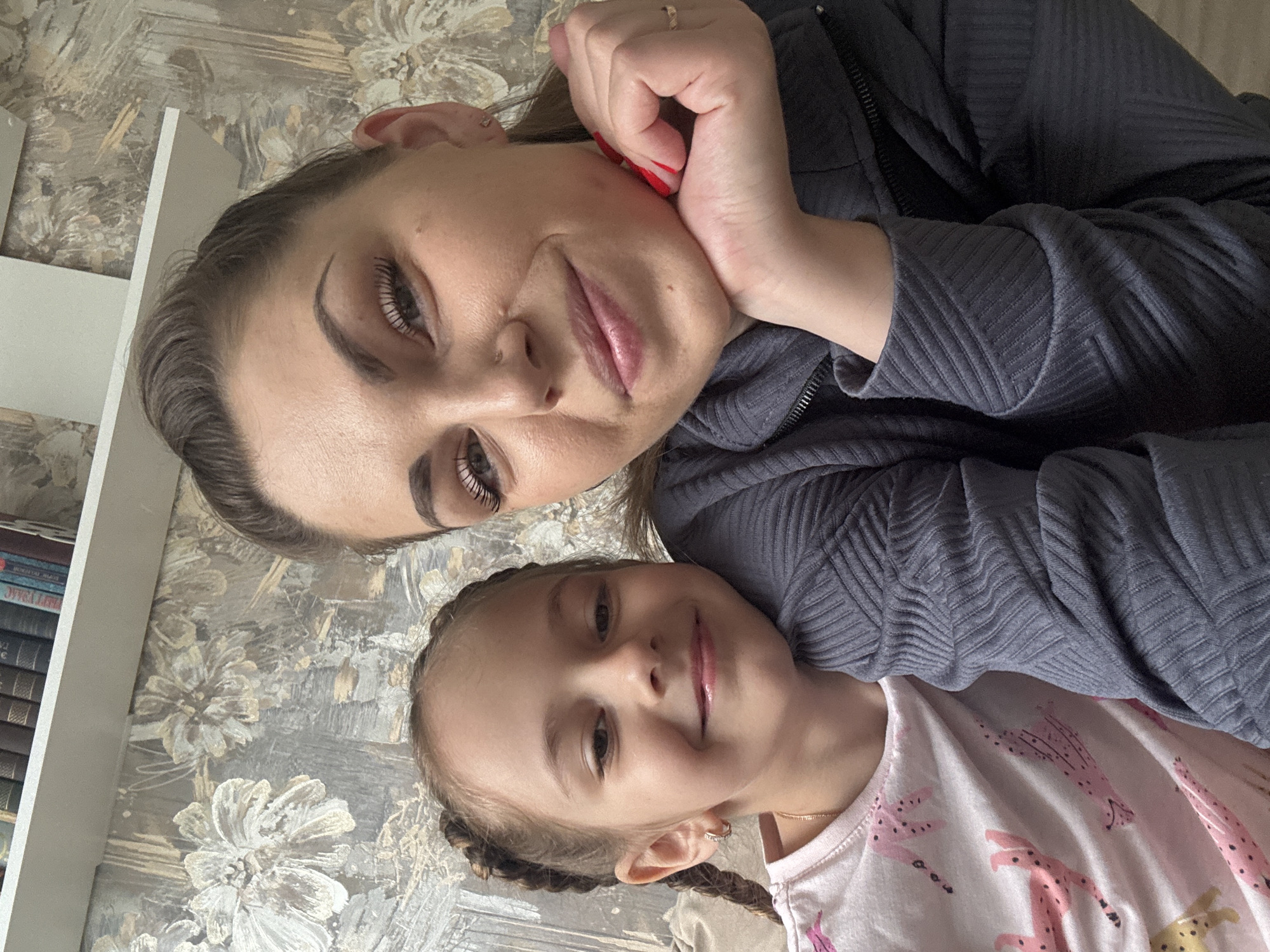 A selfie captured from an overhead perspective shows a woman and a young girl. The woman, with dark hair and pronounced eyebrows, rests her hand against her cheek. Her nails are painted red, and she's wearing a textured gray jacket. The girl, positioned below the woman, has light brown hair styled in braids. She wears a white t-shirt adorned with pink patterns. The background features a floral-patterned wall and a bookshelf along the left edge of the frame. The location is Раздолье, Russia.