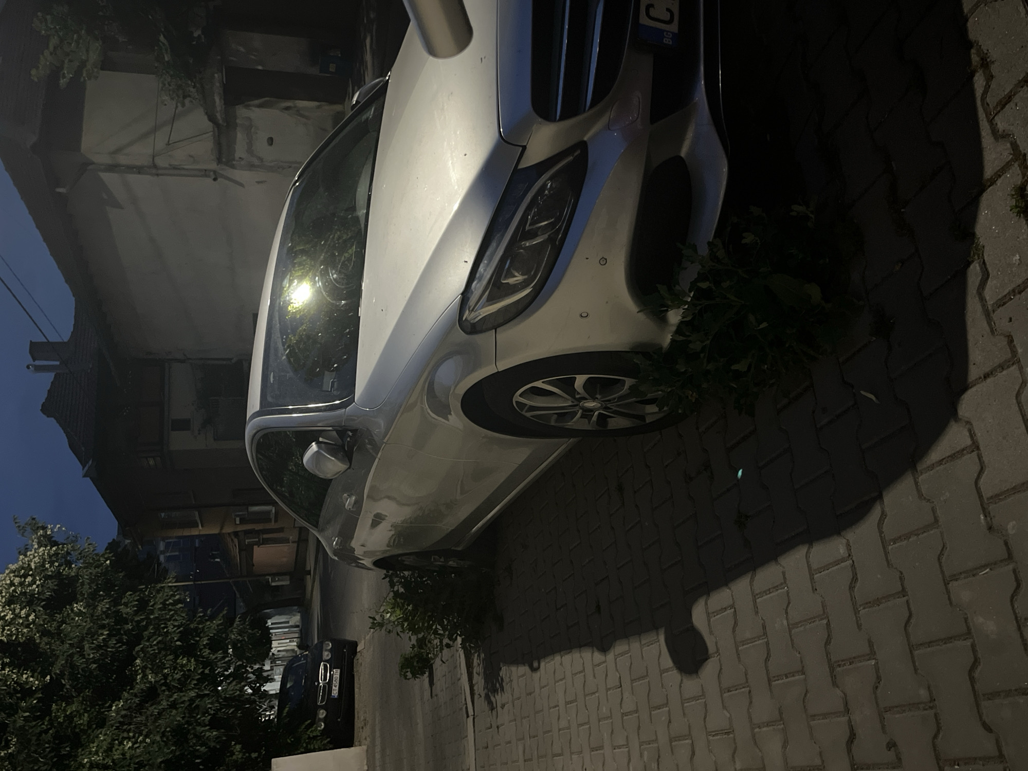The image shows a silver Mercedes-Benz car parked on a paved driveway at dusk. The car is the primary subject, with its headlights, grille, and front wheel clearly visible. To the left of the car, a dark-colored BMW is parked, its license plate "CT 4560 TB" is partially visible. A dense green tree with white blossoms is on the left side of the frame, casting a shadow. The background consists of buildings, one with a red-tiled roof, suggesting a residential area. The lighting indicates it is either early evening or late dusk, with a clear sky. The scene appears to be in Stara Zagora, Bulgaria, as indicated by the license plate format and location context. There is no discernible activity or emotion depicted; it is a static scene of parked vehicles. The overall mood is calm and quiet, characteristic of the transition from day to night.