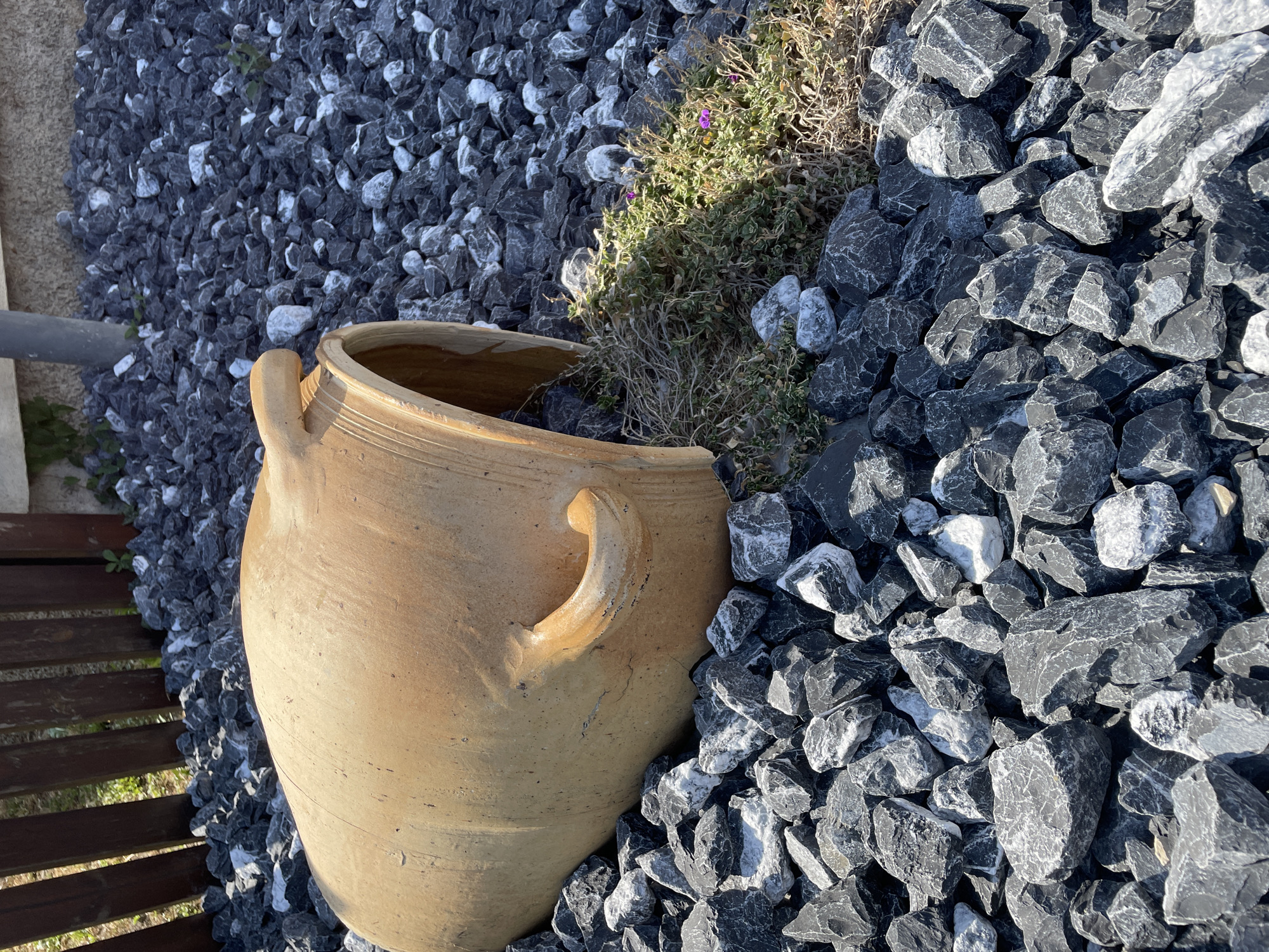 This media file is a still image taken outdoors during the daytime, likely in a garden setting. The scene is dominated by dark grey and white gravel, with a large, light brown clay pot positioned prominently in the lower left portion of the frame. The pot has two handles and appears aged, with visible cracks and texture. To the left of the pot, a section of a wooden deck or fence is visible, constructed from dark brown planks. In the upper middle of the frame, a small patch of dry-looking green and brown vegetation with tiny purple flowers is growing amongst the gravel. The sunlight casts shadows, indicating a sunny day. The overall impression is one of a decorative outdoor space, possibly a courtyard or garden path. There is no visible text in the image. The location context provided is Gerstheim, France.