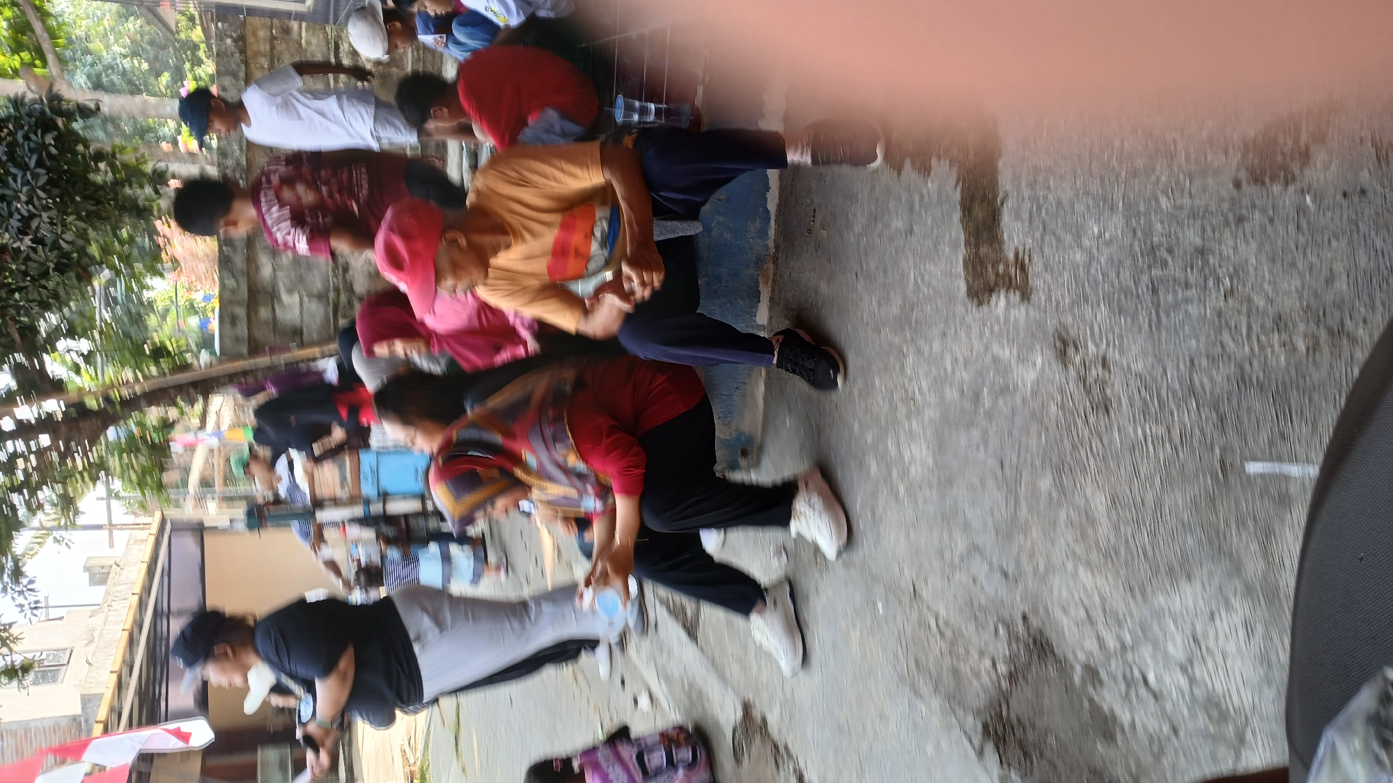 In Mendut, Indonesia, a group of people are gathered outdoors. The foreground is dominated by a worn, grey concrete surface. To the right, a blurry pink object, possibly a hand or finger, intrudes into the frame. A man in an orange t-shirt and dark blue pants sits on a blue ledge, his hands clasped in his lap. Next to him, a woman wearing a red hijab and black pants is seated, holding a clear plastic cup. She is looking to her left. Further back, more individuals are visible, some standing and others sitting. A child in a striped shirt stands near a table. In the background, a brick wall and buildings are present, along with trees providing shade. Several flags, including one with red and white stripes, are displayed. The overall scene suggests a community gathering or event.