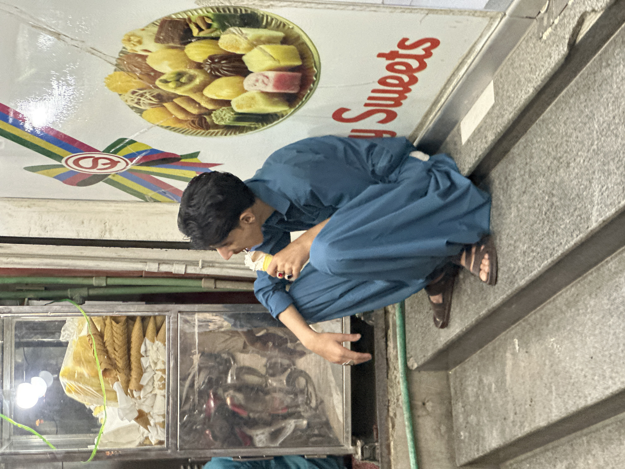 A medium, eye-level shot shows a man in Unknown, Unknown eating an ice cream cone next to a sweets shop. He is crouched with his head bent eating the ice cream, and he is wearing a long blue dress shirt with long sleeves and brown sandals. A sign above him to the right is on an angle and features a plate full of sweets, and red cursive text to the right that says "Sweets". To the man's left is a display case with many ice cream cones inside. There are green pipes overhead. To the man's right are gray steps and concrete.