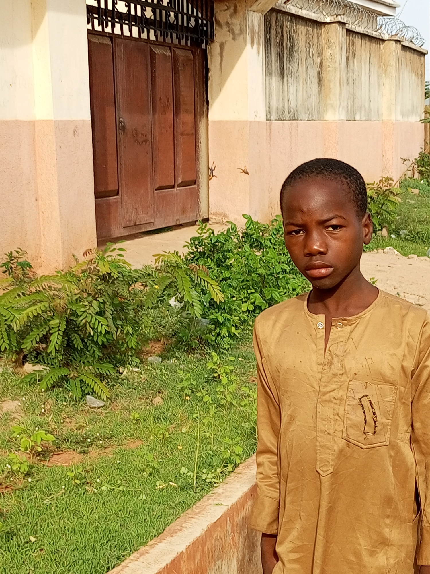 A young boy stands outdoors in front of a building with a large, weathered brown wooden door and a metal gate above it. To the right of the door, there is a wall topped with barbed wire. The boy is wearing a tan, long-sleeved tunic with two buttons at the neck and a decorative embroidery on the chest pocket. He is positioned in front of lush green foliage and grass. The ground appears to be dirt and gravel. The scene suggests a setting in Tempure, Nigeria, characterized by its warm climate and architecture.