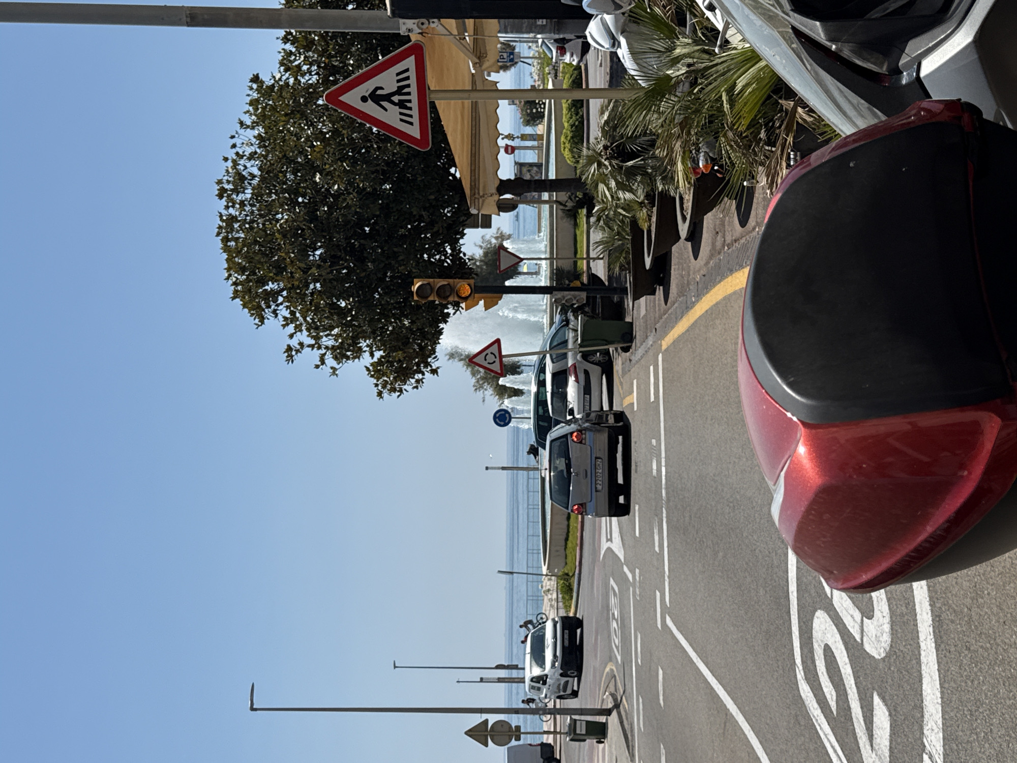The image depicts a street scene in Blanes, Spain, likely on a sunny day, with the sea visible in the background. A red scooter is parked in the foreground, partially obscuring the road and speed limit marking which shows "20". A silver car is driving in the middle distance, followed by a white car. Further back, a fountain with multiple jets is active. Traffic lights show yellow, indicating a caution or impending change. Several traffic signs are visible, including a pedestrian crossing sign (a red triangle with a black pedestrian symbol) and a circular sign with a red border and a black arrow indicating a roundabout. Potted plants and trees line the sidewalk, and a cafe umbrella is visible. The overall impression is of a coastal town with moderate traffic and pedestrian activity.