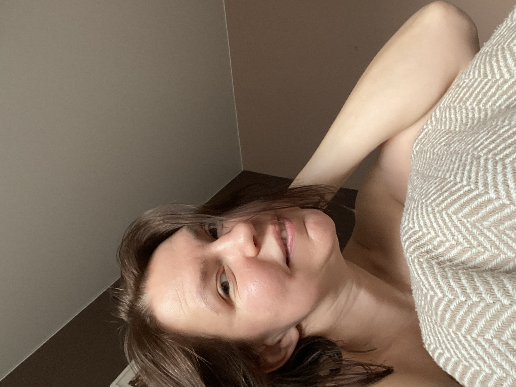 The image is a close-up, angled selfie of an adult female captured indoors. The woman, with medium-length dark brown hair, is lying down and looking towards the camera with a gentle, closed-mouth smile. Her skin is fair, and she has a natural appearance, seemingly without make-up. Her body is partially covered by a light-colored blanket or throw, which features a distinct herringbone or chevron weave pattern in beige and off-white tones.
The background consists of plain, muted-toned walls, forming a visible corner; one wall is a light grey-brown, and the other is a darker brown. The lighting appears to be natural, suggesting the photo was taken during the daytime. The overall scene conveys a sense of relaxation and comfort, implying the woman is resting on a bed or couch. No specific location cues like landmarks or unique architectural features are visible to indicate Chaikovsky, Russia, nor is any text discernible in the image.