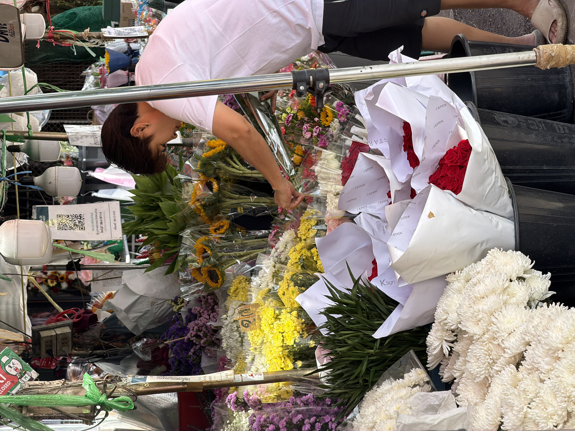 A person in a white t-shirt and black shorts is arranging flowers at a market stall. The stall is filled with various types of flowers, including sunflowers, chrysanthemums, and roses, all wrapped in plastic or paper. The person's left hand is reaching towards a bouquet of mixed flowers. In the background, there are more flowers, a QR code displayed on a sign, and overhead lighting. The scene appears to be outdoors, possibly in a street market in Chiang Mai City Municipality, Thailand, given the visual cues.