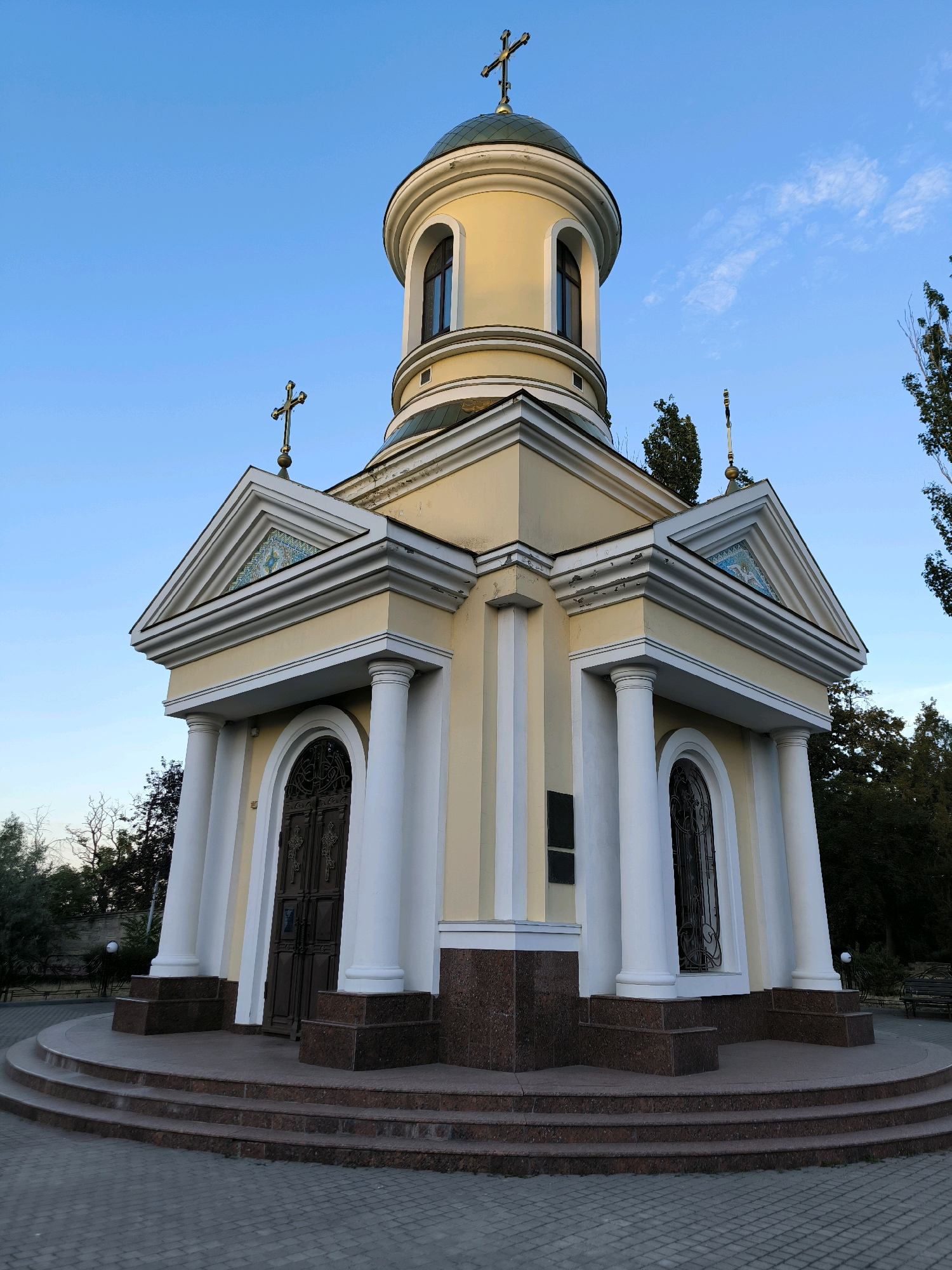 This is a photograph of the Chapel of Saint Nicholas, located in the city of Mykolaiv, Ukraine. The chapel is a small, yellow building with a domed roof topped with a cross. It has white columns, arched windows, and a large wooden door. The building is situated on a circular platform with several steps leading up to the entrance. The sky is clear and blue, with a few wispy clouds. Trees and foliage are visible in the background. The overall scene is peaceful and serene.