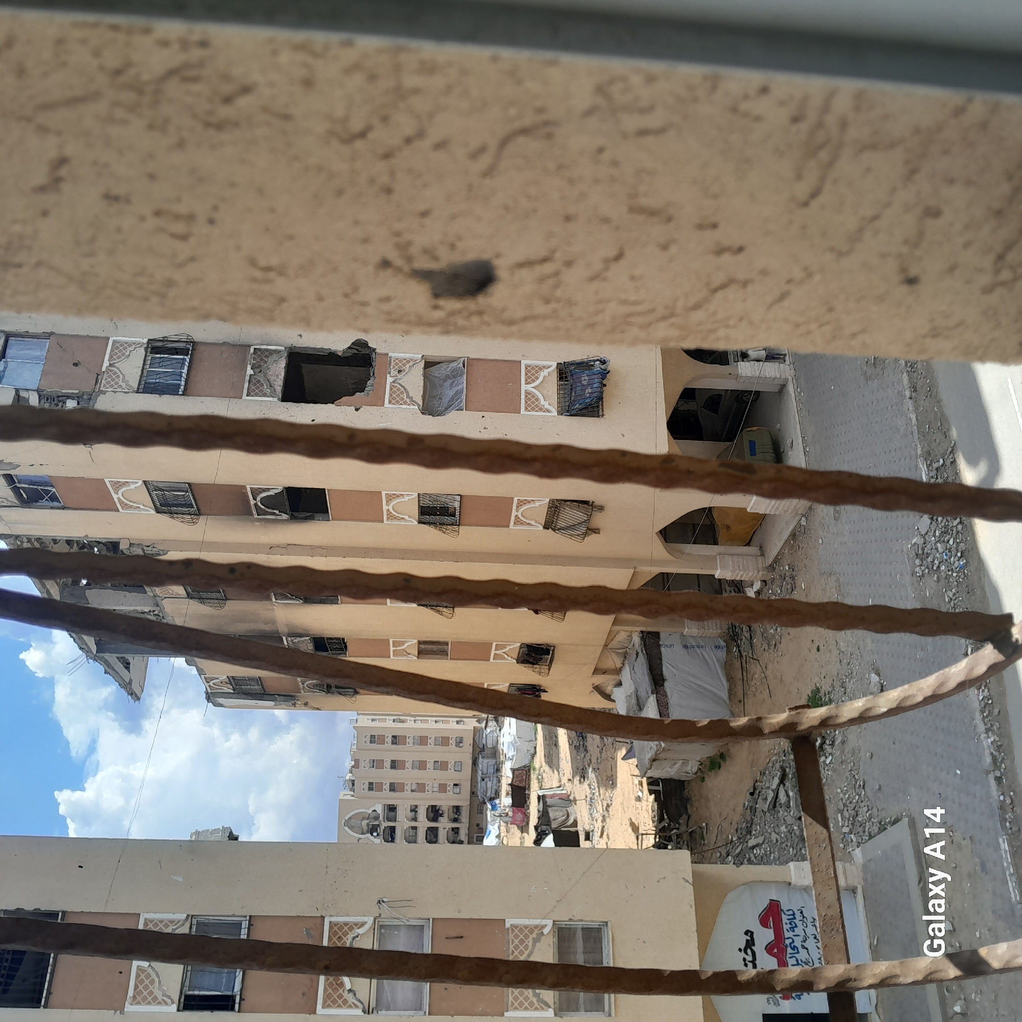 Here's a matter-of-fact description of the image:
The image shows a view from an elevated position, possibly a balcony or window, overlooking a cityscape in what appears to be an urban environment. In the foreground are twisted metal bars of a railing. The buildings in the view appear to be densely packed. Many buildings show signs of damage with missing windows and structural decay. In the background, taller apartment buildings are visible. The sky is visible in parts of the image with blue sky and clouds. In the lower right corner of the image, a sign with Arabic script is visible, and the camera details "Galaxy A14" are printed. Debris and rubble are visible in the open spaces between buildings.