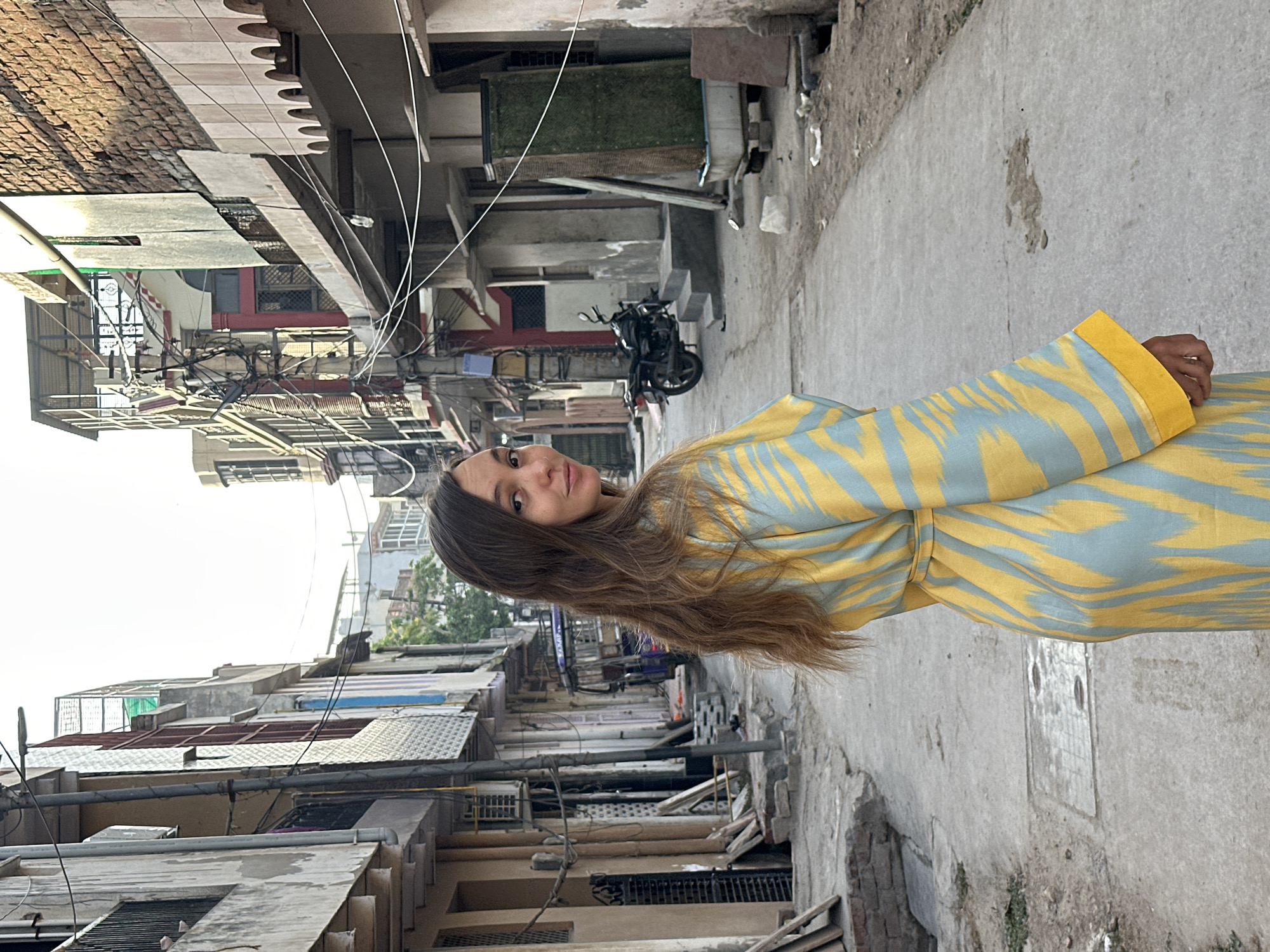 A young woman stands in a narrow street in Mathura, India. She is positioned to the right of the frame, wearing a light-blue and yellow-striped garment with yellow cuffs. Her long brown hair flows down her shoulder. The street, made of concrete, is flanked by aged buildings. The buildings on both sides are a mix of residential and commercial structures, with visible signs of wear and tear. Utility wires crisscross overhead, adding to the cluttered urban atmosphere. A parked motorcycle can be seen further down the street. The architecture features a mix of brick and concrete, with windows and balconies lining the facades. The overall environment suggests an older, densely populated urban area.