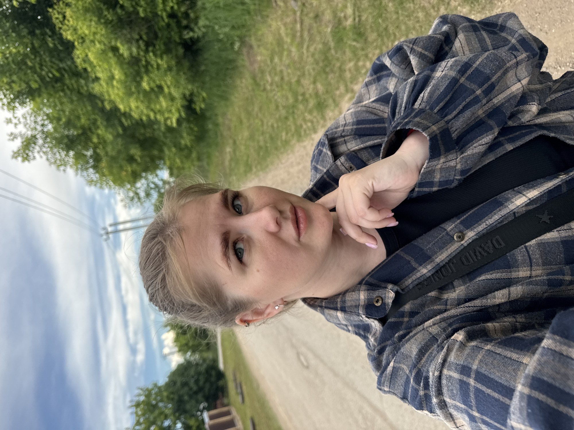The media file is a portrait-oriented selfie of a young woman outdoors. She is wearing a blue and tan plaid flannel shirt over a black top, with a black strap visible across her chest that appears to be part of a bag. Her blonde hair is pulled back, and she has on light makeup, including mascara and lipstick. She is touching her chin with her right hand. The background shows a path or dirt road bordered by grass and trees, with a cloudy sky visible above. Power lines are also present in the sky. The lighting suggests it is daytime, possibly late afternoon. The setting appears to be rural or semi-rural. Visible text on the strap reads "DAVID JONES". The overall mood seems casual and relaxed.