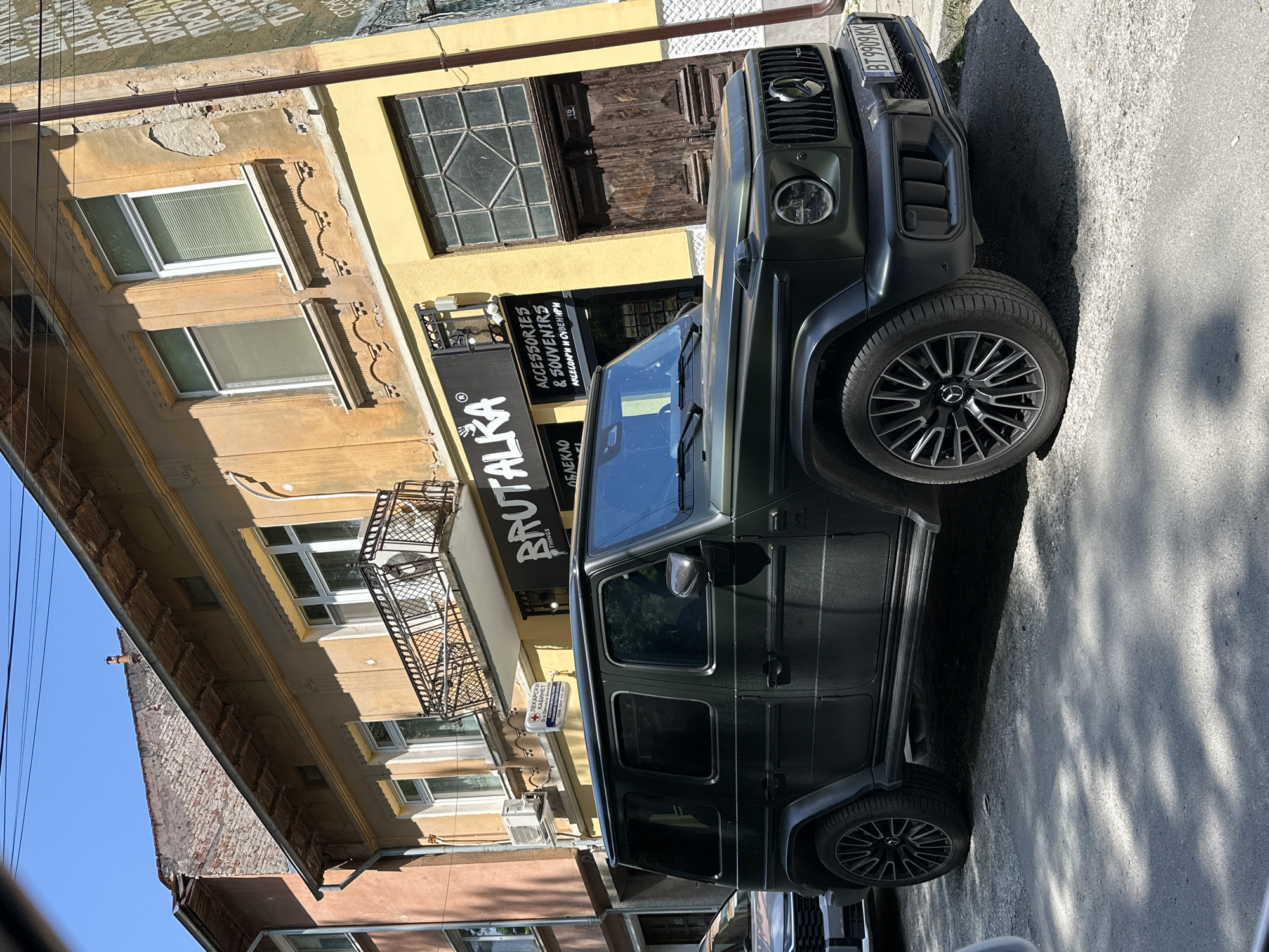 The image shows a dark green Mercedes-Benz G-Class SUV parked on a street in Veliko Tarnovo, Bulgaria. The front of the vehicle is angled towards the upper right of the frame. The car's license plate reads "BT 9909KK".
In the background, there are buildings of different colors, primarily yellow and brown. A sign for "BRUTALKA - ACCESSORIES & SOUVENIRS" is visible on the front of one of the buildings, next to a sign that reads "ЛЕКАРСКИ КАБИНЕТ". The buildings appear to have traditional architectural details, including windows and balconies. A door with the number "15" above it is also visible.
The street is paved and shows signs of wear and sunlight. The lighting indicates it is daytime. There are no people visible in the immediate vicinity of the car.