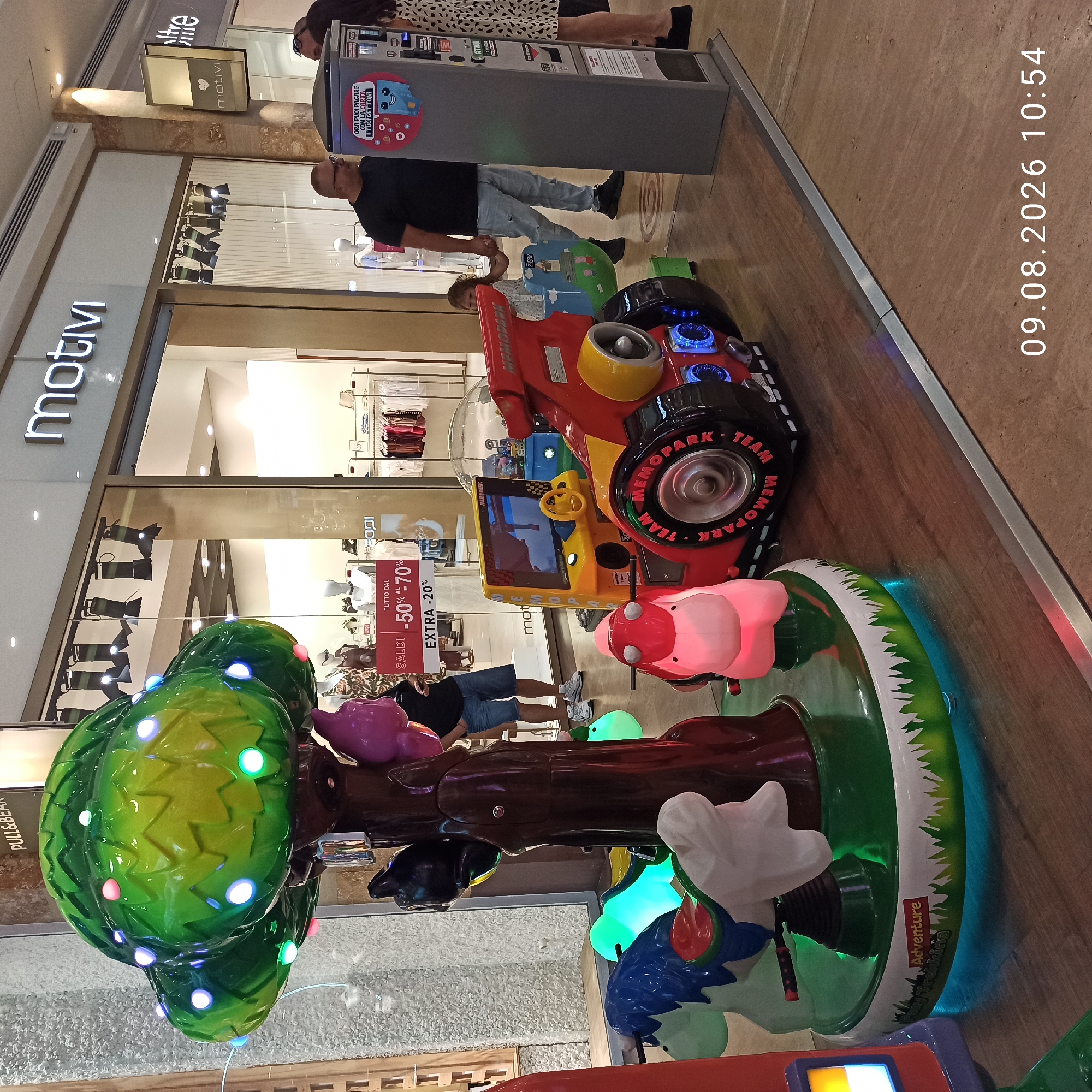 The image depicts a shopping mall interior in Molfetta, Italy. In the foreground, there is a children's carousel with a large, green, illuminated tree at its center, adorned with colorful lights. Several small, stylized animal figures, appearing to be a horse and a pink creature, are attached to the carousel base. To the right of the carousel, there is a red and yellow ride-on toy vehicle with the inscription "TEAM MEMOPARK". Adjacent to this is a grey payment machine displaying information about payment methods. In the background, store windows are visible, with one displaying a "SALDI -50% AL -70% EXTRA -20%" sign, indicating a sale. People are present in the scene: a man in a black t-shirt and blue jeans stands near the payment machine, a young girl holding his hand is looking at the ride-on toy, and a woman in a patterned dress is also near the payment machine. The overall environment appears to be clean and well-lit, consistent with a modern shopping center. A timestamp "09.08.2026 10:54" is overlaid on the bottom right of the image.