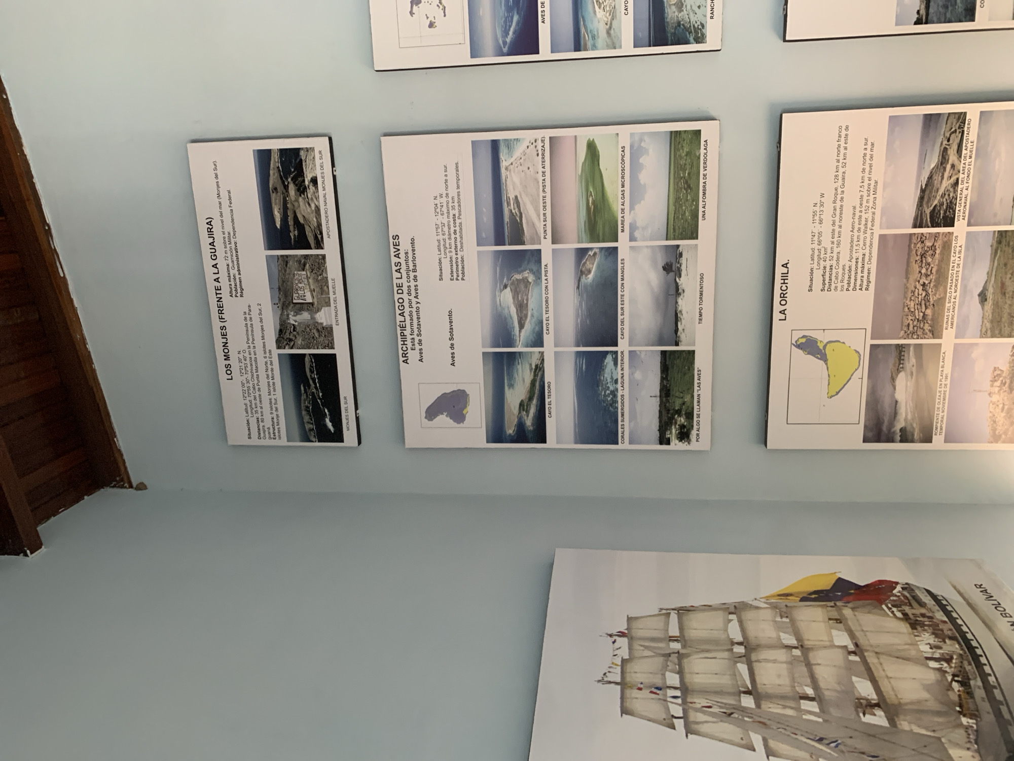 The image shows a light blue wall in what appears to be an indoor exhibit. The wall is adorned with several framed informational posters and a large framed painting of a tall ship.
The topmost poster, titled "LOS MONJES (FRENTE A LA GUAJIRA)", displays information about a group of islands, including their coordinates, distances, maximum altitude, and administrative status. It is accompanied by three landscape photographs of the islands and a caption "MONJES DEL SUR".
Below this poster is another, titled "ARCHIPIÉLAGO DE LAS AVES". This poster details an archipelago formed by two sets of islands, "Aves de Sotavento" and "Aves de Bartolome". It includes similar informational text regarding location, distances, population, dimensions, and maximum altitude. This poster is illustrated with a map of the archipelago and six photographs showcasing various aspects of the islands, such as beaches, lagoons, and marine life.
To the right of these two posters, partially visible, is another poster with similar formatting, showing a map and photographs related to islands.
Below the "ARCHIPIÉLAGO DE LAS AVES" poster, the bottom poster visible is titled "LA ORCHILA". It provides details about this island, including its situation, distances to other locations, surface area, population, dimensions, maximum altitude, and regime. This poster is also accompanied by a map and five photographs depicting different scenes on the island, including ruins, aerial views, and a beach with waves.
To the left of the posters, a large framed painting of a historical sailing ship, identified by the text "GRAN BOLÍVAR" at the bottom, is displayed. The ship is adorned with numerous flags.
The image is taken from a slightly elevated angle, looking upwards towards the top of the wall and the ceiling, which is made of wood planks. The lighting appears to be ambient room lighting, casting soft shadows. The scene does not contain any people. The posters and painting are professionally mounted on the wall. The context suggests an exhibition or display related to Venezuelan geography and history, potentially in a museum or public building. The city of Boca del Río, Venezuela is not directly identifiable from the visual elements within the image itself.