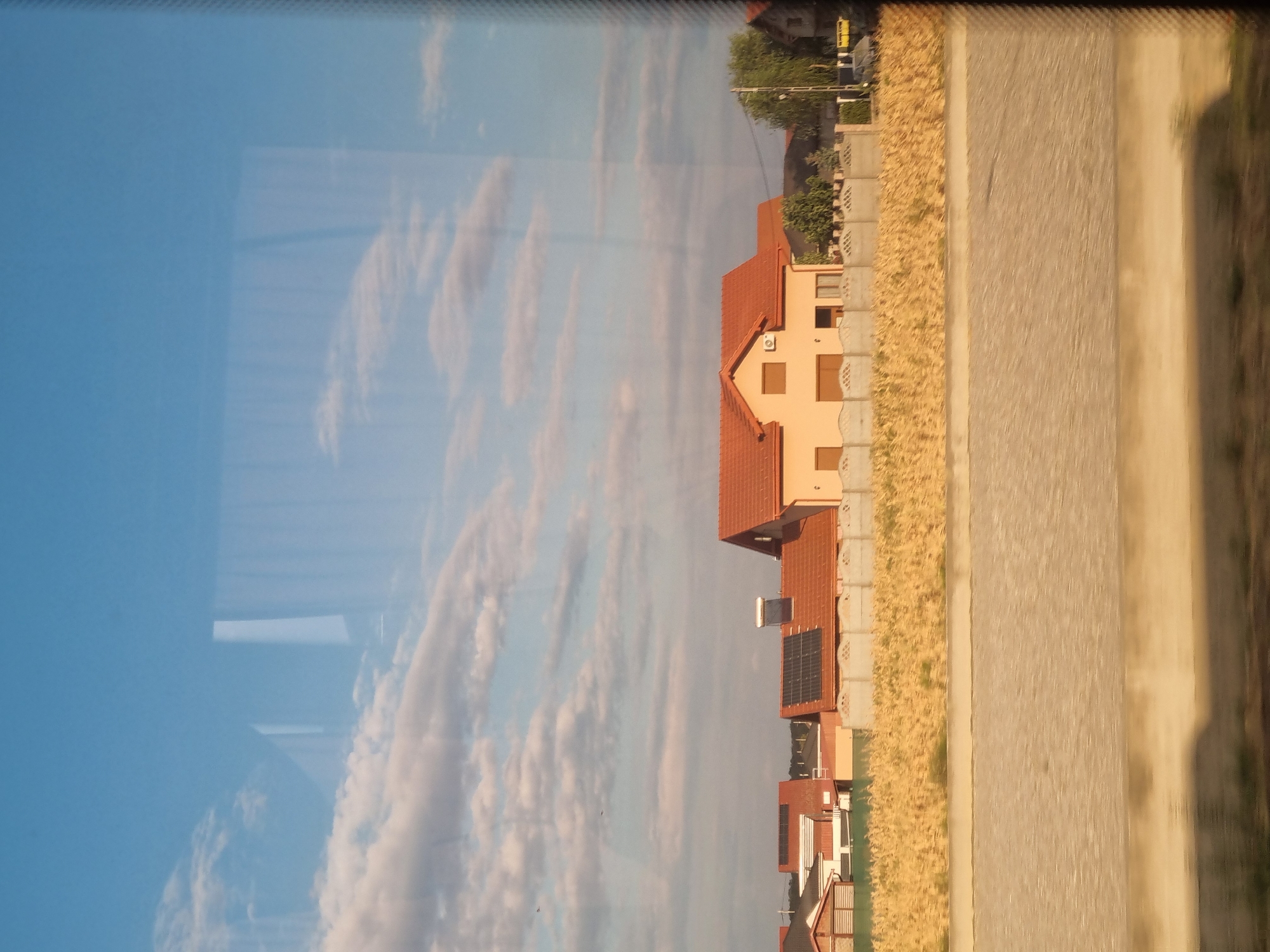 The image captures a rural landscape under a bright blue sky with scattered white clouds. In the foreground, a light brown, paved surface with a distinct pattern runs horizontally across the lower half of the frame. Above this, a field of dry, golden vegetation stretches out, creating a textured, yellowish band.
Behind the field, a row of houses with red-tiled roofs is visible. The most prominent house is a light peach-colored building with a prominent red roof. It has several windows and an air conditioning unit mounted on the wall. Some of the other houses in the background also have red roofs and appear to be of a similar architectural style. Several of these houses are equipped with solar panels on their roofs. A concrete fence separates the houses from the field.
To the right of the houses, there is a cluster of green trees, and a street light pole stands tall. The overall impression is of a serene residential area in a rural setting, likely in the city of Nojorid, Romania, given the context. The image appears to have been taken through a window, as evidenced by reflections on the surface.
