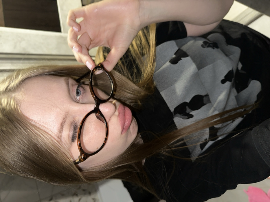 A young female individual with long, straight, light brown hair, light blue eyes, and fair skin is depicted in a close-up, indoor setting in the city of Chaikovsky, Russia. She is wearing a black long-sleeved shirt under a black short-sleeved t-shirt adorned with a grey and black checkered pattern featuring dark silhouettes of people. Her right eye is closed, her left eye is open, and her lips are pursed into a pout. She holds a pair of tortoise-shell framed glasses with her right hand, positioning them below her left eye. The background is indistinct, showing a white tiled surface on the left and a light-colored vertical structure on the right.