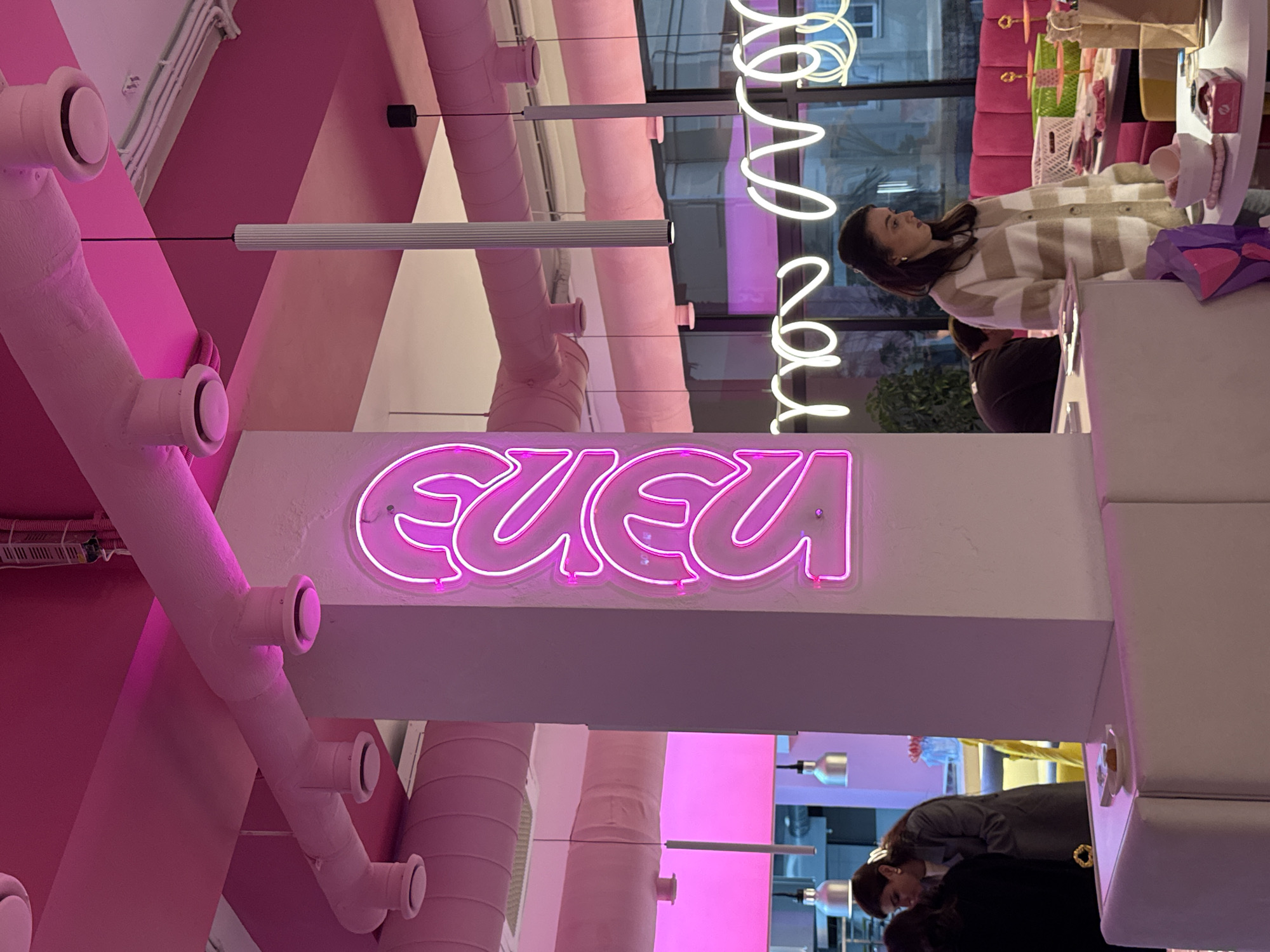 The image shows the interior of a shop in Sochi, Russia, featuring pink decor and the name "EUEU" in neon pink letters. Inside, there are at least two people present: a woman in a striped sweater standing and potentially shopping, and a second person in a black shirt. The shop appears to sell a variety of items, some visible on shelves and displays. Pink ventilation pipes are a prominent feature on the ceiling, along with additional neon lighting.