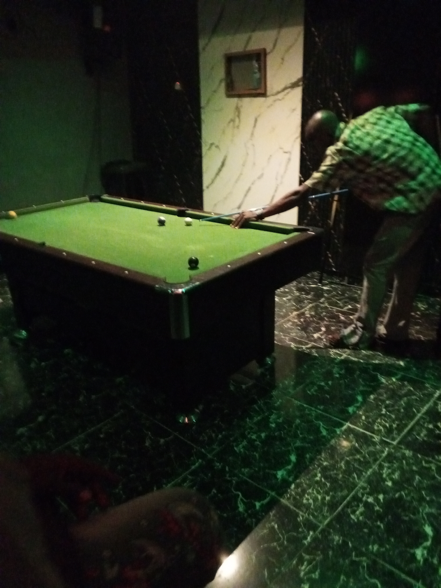 In an indoor setting, a person is playing billiards on a green pool table. The room appears to be dimly lit, with green lighting casting reflections on the marble-patterned floor. The person is wearing a green and white checkered shirt and light-colored pants. They are positioned behind the pool table, holding a cue stick and aiming to strike a ball. Several billiard balls are visible on the table, including a white cue ball and a darker ball. The background features a marble-patterned wall with a small framed picture. To the left of the pool table, a dark, possibly upholstered, object is visible. The overall scene suggests a recreational environment.