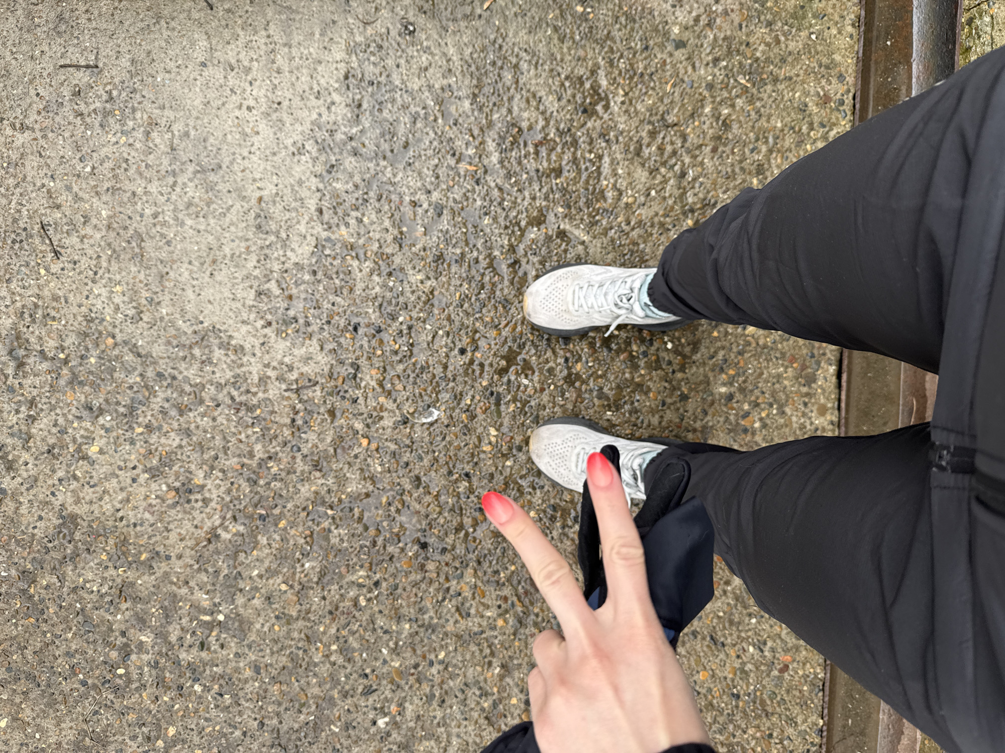 An overhead shot captures a person's lower body and one hand against a backdrop of wet, textured concrete, located in Chaikovsky, Russia. The person is wearing black pants and light-colored athletic shoes with light blue accents. The left foot and leg are partially visible on the right side of the frame, while the right foot and leg extend towards the bottom right. The ground beneath them is a coarse concrete mix, embedded with numerous small pebbles of various shades of gray, brown, and white. Patches of the concrete appear damp and glistening, suggesting recent rain or wet conditions.
In the bottom right portion of the image, the person's right hand is prominently displayed, making a "peace" sign (V-sign) with the index and middle fingers raised. The fingernails on these fingers, and possibly others, are painted with a striking two-tone gradient, transitioning from a light, natural color at the base to a vibrant, bright red at the tips. A darker, possibly metallic or worn concrete edge, runs along the far right side of the image, separating the paved area from what appears to be another surface. The lighting is natural, indicating it is daytime. There is no visible text in the image.