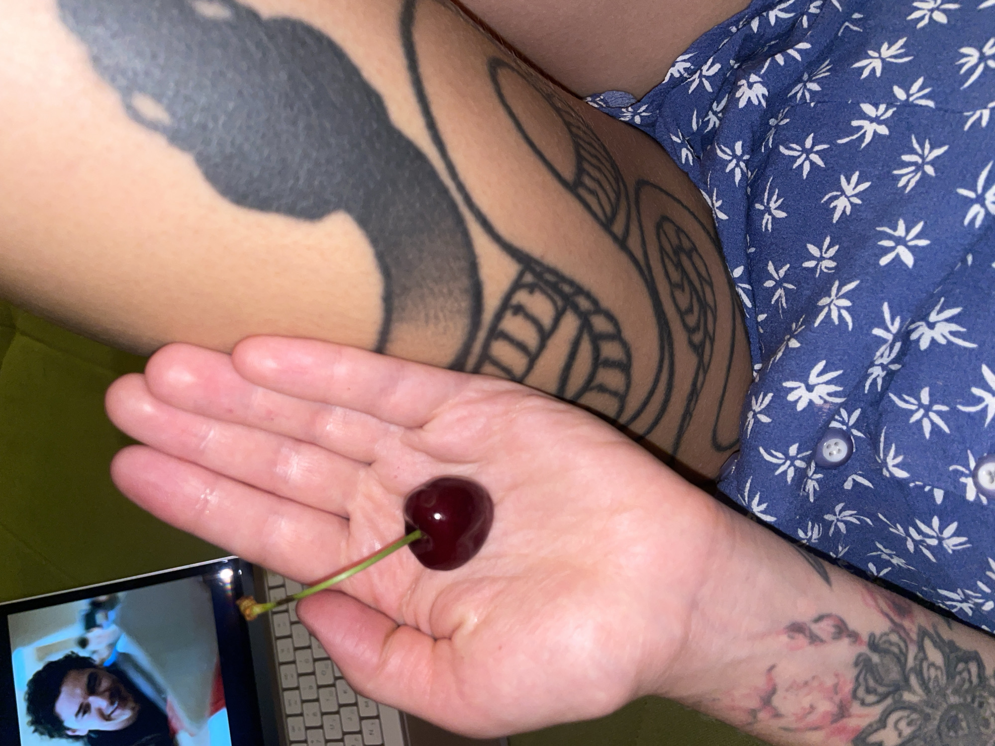 A person's hand is holding a single ripe cherry with a green stem. The hand is in the foreground, positioned in the lower left quadrant of the image, with the cherry resting on the palm. The person's forearm, also visible on the left, features a tattoo. To the right of the hand, a tattooed leg is visible, displaying a large snake design. The person is wearing a blue floral patterned garment, which is in the lower portion of the image. In the upper left corner, a laptop screen is partially visible, showing a smiling man's face. The background appears to be a textured surface, possibly upholstery. The lighting is dim, with the primary light source seemingly originating from the laptop screen. There is no indication of the city of Boreti, Montenegro in this image.