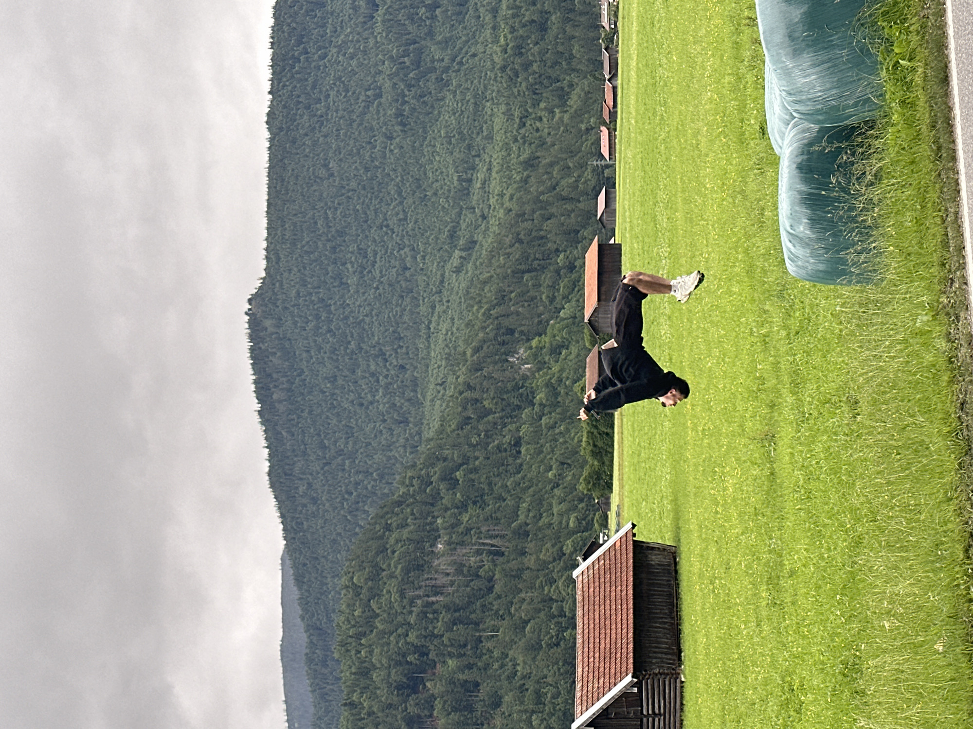 An individual is captured mid-flip in a grassy field in Garmisch-Partenkirchen, Germany. The person, dressed in dark athletic wear, is performing a backflip with their body inverted and legs extended. Behind them, a dark wooden barn with a red-tiled roof stands to the left. Further in the background, several other similar structures are visible. The field is lush green, dotted with small yellow wildflowers. Towering, densely forested mountains form the backdrop, under a heavily overcast, grey sky. In the foreground, three large, round bales of hay, wrapped in green plastic, are positioned on the right side of the frame. A grey road borders the bottom edge of the image.