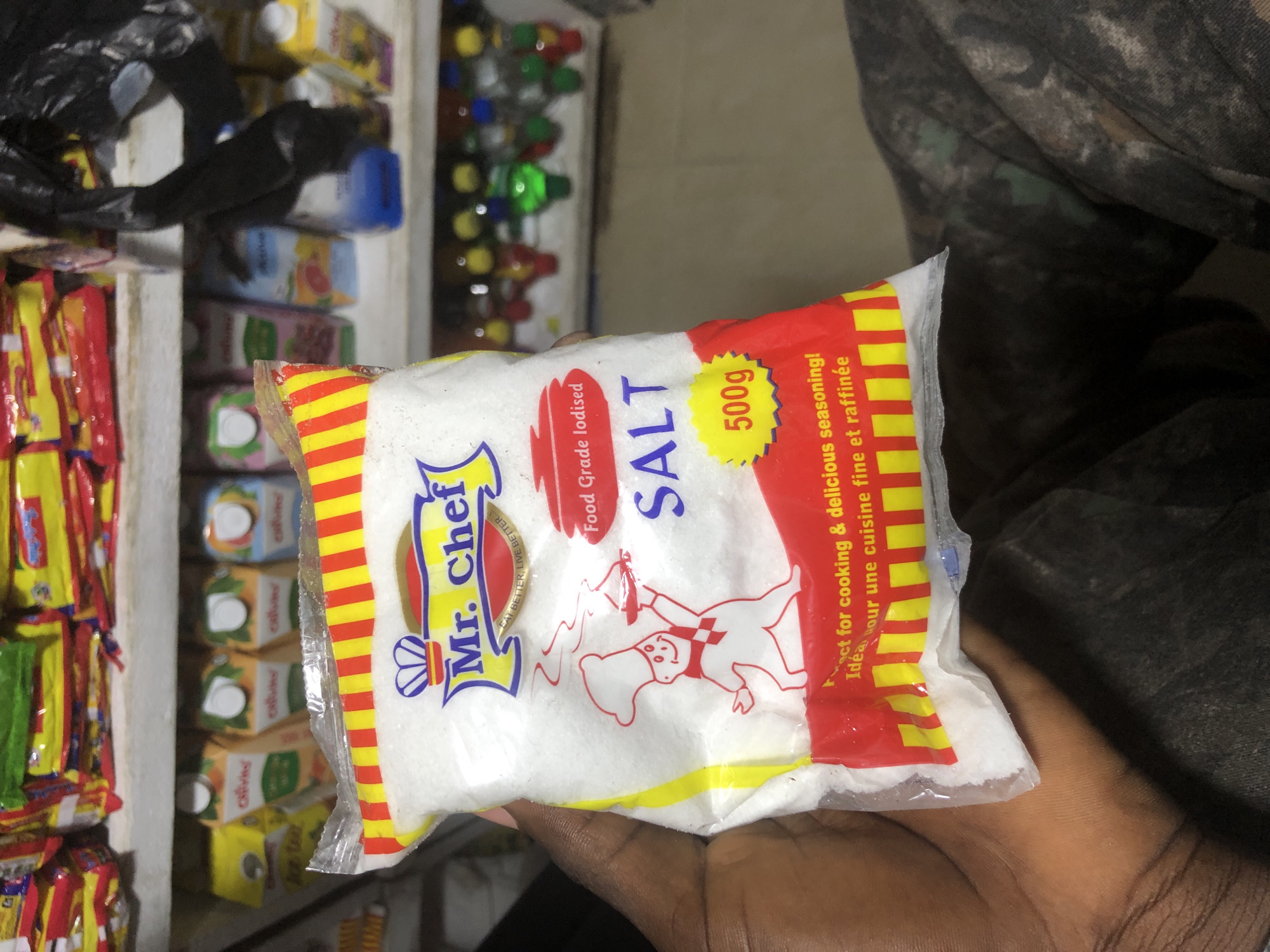 The image shows a close-up of a person's hand holding a bag of Mr. Chef salt. The bag is mostly white with red and yellow stripes at the top. There is a cartoon drawing of a chef on the front of the bag, and the words "Mr. Chef" are prominently displayed. Below the chef, the words "SALT" are written in large blue letters, and to the right of that, a yellow oval contains "500g". Text below the salt states "Perfect for cooking & delicious seasoning!" and "Idéal pour une cuisine fine et raffinée".
The background of the image shows shelves stocked with various products, including what appear to be juice cartons and bottles of beverages. The overall impression is of a small shop or market stall. The presence of the "Mr. Chef" brand of salt and the general appearance of the goods suggest a retail environment, potentially in a Nigerian city like Ado, where this brand is common.