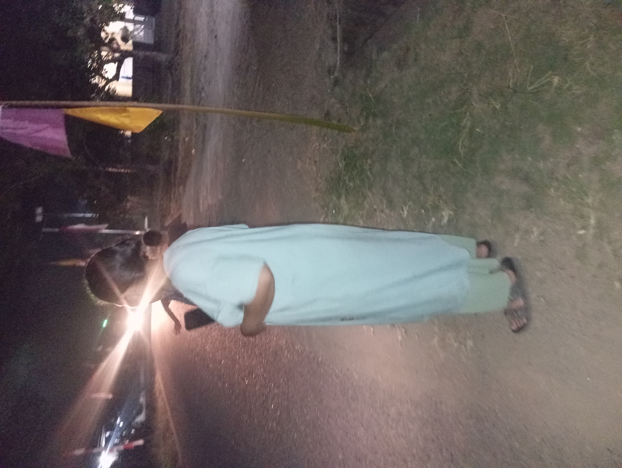 The image depicts a dimly lit outdoor scene, likely at night, on a dirt road in Rejosari, Indonesia. A woman, seen from the side and back, stands on the dirt road facing away from the camera. She is wearing a light blue, long-sleeved tunic over light green pants and dark sandals. Her hair is dark and pulled back. To her left, a bright light source, possibly from a vehicle on a paved road further in the background, illuminates the scene and creates a lens flare. In the right portion of the image, a thin wooden pole stands upright, with a purple flag and a yellow flag attached to it, blowing in the wind. Trees with visible foliage are present in the background, and patches of grass and dirt make up the ground. There is another blurred figure in the far background.
