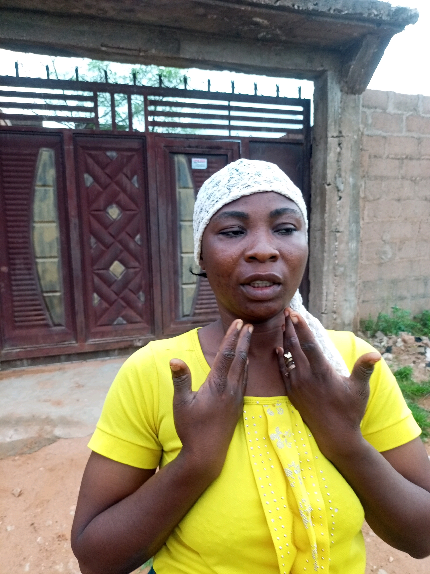 A dark-skinned woman with a white headscarf and a yellow shirt is pictured outdoors. She is holding her hands up to her neck and chest. Her shirt is adorned with small white dots and a decorative pattern. Behind her, there is a large, dark red double gate with decorative panels and a metal fence above it. To the right of the gate, a textured brick wall extends upwards. The ground is a mix of dirt and gravel. The overall setting suggests a residential area in Tempure, Nigeria.