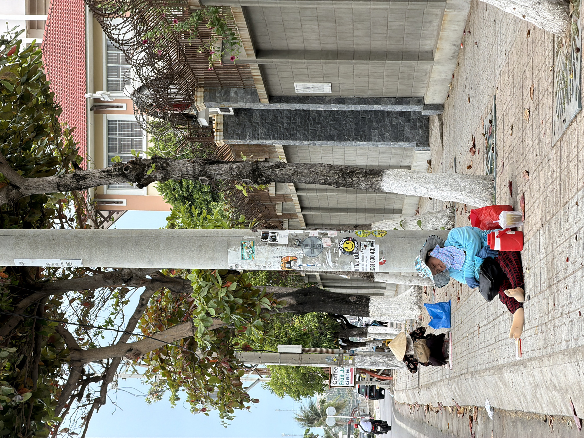The image shows a street scene in Bac Nha Trang Ward, Vietnam. A woman is reclining on the sidewalk, wearing a light blue shirt, dark pants, and a patterned hat. Red and blue bags and a red container are nearby. An unidentifiable individual wearing a traditional conical hat and other bags is also on the sidewalk, further down the street. A light gray utility pole is located adjacent to the reclining woman, covered with stickers and graffiti. Trees line the street, with their trunks painted white. A building is visible in the background, featuring a red-tiled roof, windows with bars, and a fence topped with barbed wire. The sidewalk is paved with a patterned material and scattered with leaves. Motorbikes are driving on the street. A sign for "Coffee Fruit Ju" is visible on the pole.