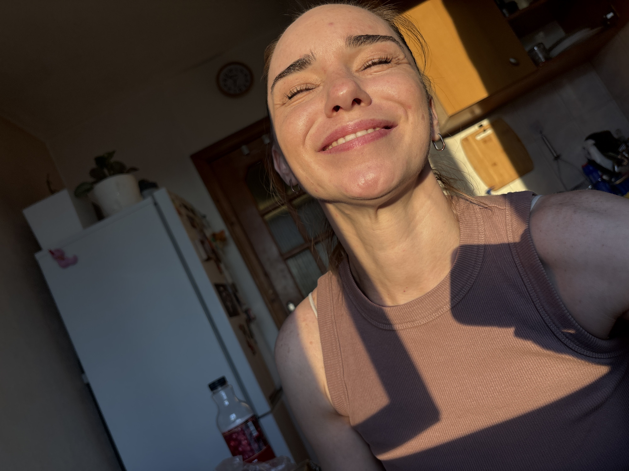 A close-up, selfie-style shot features a woman with her eyes closed and a broad smile. The lighting, which appears to be natural and golden from the left, casts strong shadows across her face and the ribbed, pink tank top she is wearing. She has on small hoop earrings. In the background, the scene is a domestic interior, possibly a kitchen, with a white refrigerator on the left, a wooden cabinet with shelves above, and a door frame. A plant in a white pot sits on top of the refrigerator. The overall impression is of a casual, candid moment captured in what seems to be a home environment. The location is identified as Unknown, Russia.