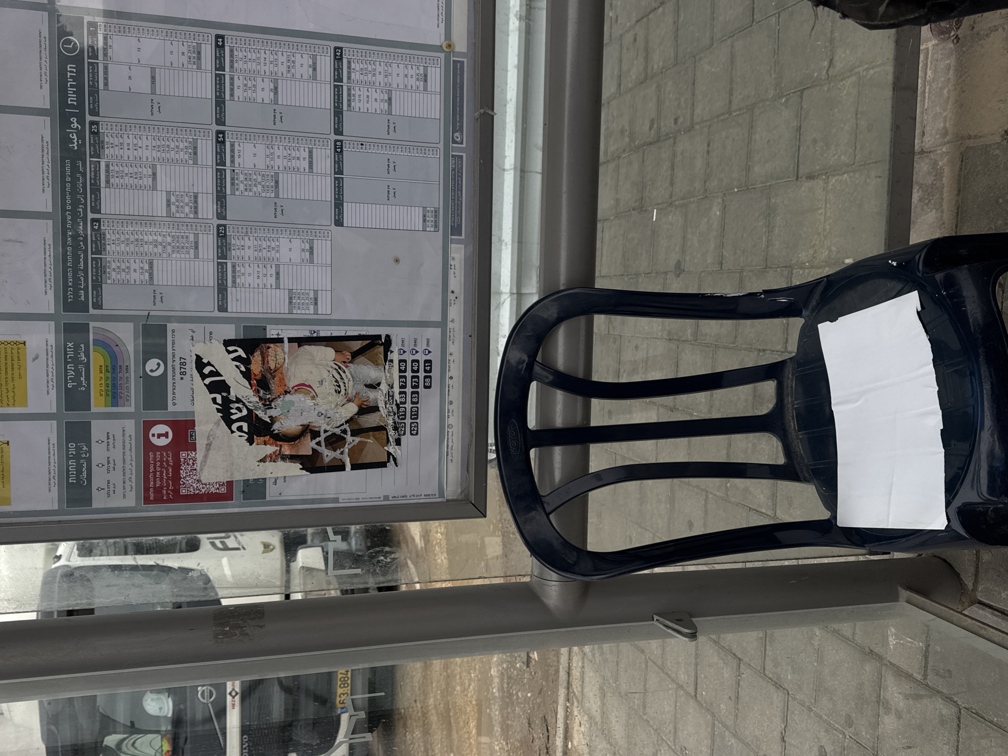 The image captures a bus stop shelter situated on a paved sidewalk in Tel-Aviv, Israel. The shelter consists of a grey metallic frame and transparent panels. A large display board on the right presents bus schedules in Hebrew for routes including 25, 42, 1, 125, 54, 44, 418, and 142. To its left, another board displays general public transport information in Hebrew, including "City Areas" and "Types of Stations," with a QR code. A prominently affixed, heavily damaged and partially torn poster is layered over other signage; it features a distressed image of a child with a stylized Star of David symbol superimposed. Below this poster, Hebrew text and numbers (e.g., 425, 119, 83, 73, 40, 88, 41) are visible.
A dark blue plastic chair, marked with the brand "MOTOR," is positioned inside the shelter, with a crumpled piece of white paper or tissue resting on its seat. Through the transparent panels, a large white commercial vehicle, likely a bus, displaying "FL" on its side and a partially visible license plate "63-884," is seen on a dirt-covered road in the background. The ground within the bus stop area is paved with grey interlocking concrete pavers. The lower leg and foot of an individual, wearing dark trousers and a shoe, are partially visible on the far right of the frame, indicating a person standing nearby.