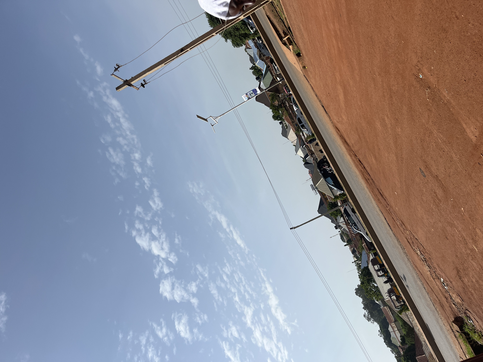 The image depicts a street scene in Bukuru, Nigeria, under a clear blue sky with scattered white clouds. The foreground is dominated by a reddish-brown dirt road or area, with some sparse green grass at the bottom left.
A paved road runs diagonally across the mid-ground, with vehicles visible parked and moving. These include what appear to be a white van or SUV, a dark-colored car, and at least two auto-rickshaws or "keke napeps" (a common mode of transport in Nigeria), one yellow and one with a darker hue. There are also utility poles with wires running across the scene. One prominent wooden utility pole stands in the right half of the image, angling upwards, and a modern lamppost with a solar panel is visible further down the road.
Buildings with various roof colors, including gray, green, and brown, line the far side of the road, suggesting a residential or commercial area. Trees are interspersed among the buildings.
In the far right of the image, the face of a person wearing a white head covering is partially visible, looking towards the scene. The overall impression is of a typical urban or semi-urban environment in Nigeria during daylight hours. The image is taken from a slightly elevated perspective and is tilted.