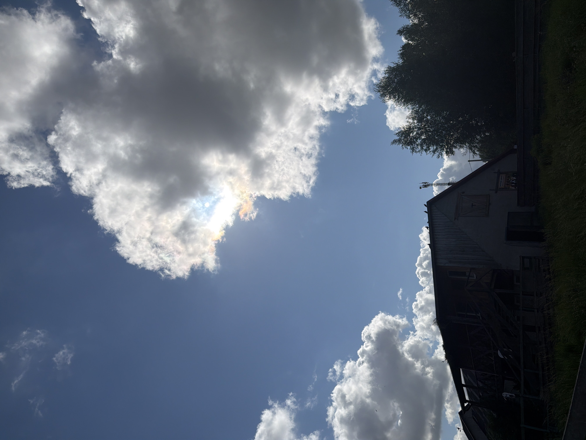 The image depicts a rural scene with a large, dilapidated structure in the foreground, possibly a barn or an old industrial building. The structure is silhouetted against a bright sky filled with dramatic clouds. Sunlight breaks through a large cloud formation, creating a halo effect. To the right of the structure, a dense line of trees is visible, also in silhouette. The lower portion of the image shows overgrown grass and a dark horizontal line, possibly a fence or a road. The presence of a cell tower in the background suggests proximity to a populated area. The image appears to have been taken in the city of Konstantinovka, Russia, indicated by the metadata. The overall mood is somewhat overcast and potentially melancholic, with the decaying structure contrasting with the dynamic sky.