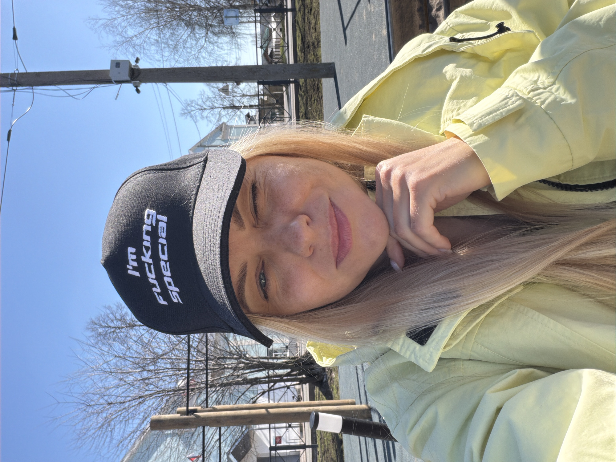 A woman with blonde hair and wearing a black cap with white text "I'm FUCKING SPECIAL" and a yellow jacket is lying down outdoors on what appears to be a park bench or similar structure. Her head is resting on her hand, and she is looking at the camera with a slight smile. The scene is set outdoors, likely in daylight, with bare trees and other structures such as poles and buildings visible in the background. Based on these features, it is not possible to identify the precise location as Votkinsk, Russia.