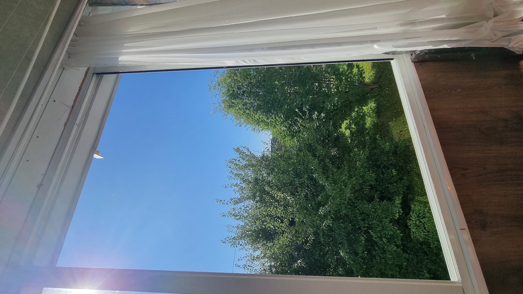 A clear, bright, sunny day is viewed through a window looking out onto lush green trees. The sky is a vibrant, cloudless blue. To the right of the frame, a sheer white curtain hangs, partially obscuring the view. The window frame is white, and below the window, a wooden paneling is visible. The scene depicts an outdoor environment, likely a residential area, given the presence of trees and the window with a curtain. The sun is shining brightly on the left side of the image, casting a lens flare.