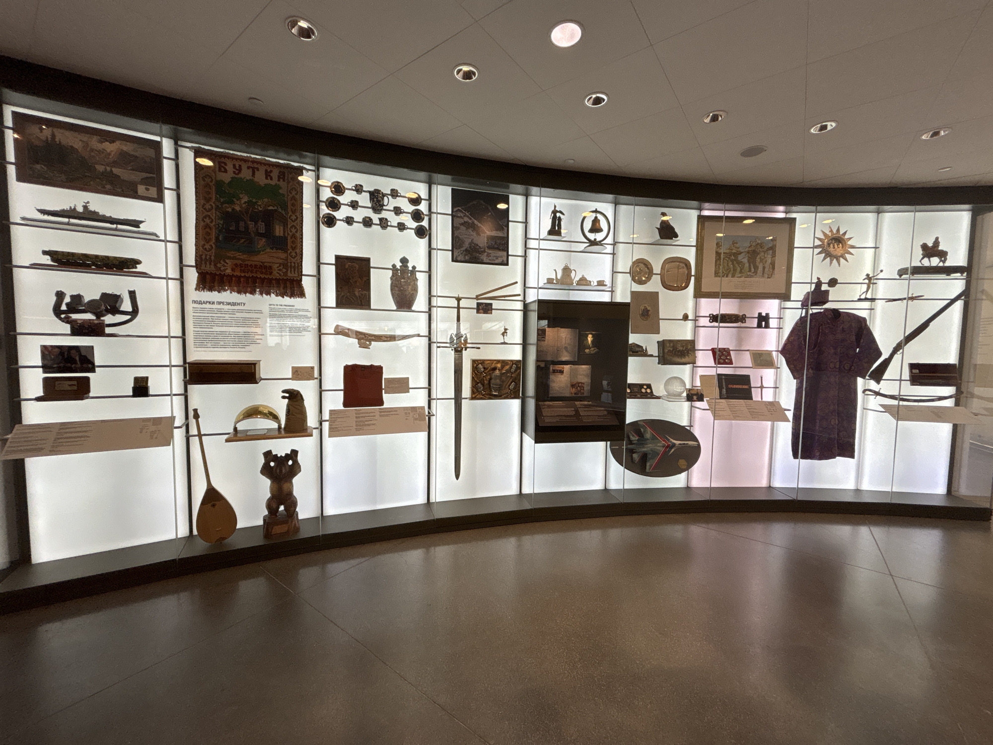 The image depicts a well-lit, modern museum exhibit inside the Boris Yeltsin Presidential Center in Yekaterinburg, Russia. The scene features a wide, curved display wall divided into numerous vertical and horizontal compartments, each backlit with white light and protected by clear panels. The floor is dark and reflective, suggesting polished concrete or stone.
The exhibit showcases a diverse collection of artifacts labeled as "GIFTS TO THE PRESIDENT" (and "ПОДАРКИ ПРЕЗИДЕНТУ" in Russian). These objects are arranged on multiple shelves within the compartments:
* On the left, a framed landscape tapestry hangs above a detailed model of a battleship or aircraft carrier. Below it, a long, flat musical instrument (possibly an accordion or wind instrument) and an ornate metal object, possibly part of a saddle or harness, are displayed.
* A large, colorful tapestry featuring a house with the word "БУТКА" (Booth/Kiosk) and the year "1983" is prominent in the upper-middle left section. Nearby, a collection of small, dark, ornate cups or bowls is arranged on shelves.
* Further along the curved wall, items include a large, decorative vase, a framed mountain landscape picture, a small bronze statuette, and a bell on a stand.
* A delicate porcelain tea set (teapot and cups) is displayed alongside various decorative plates and a sun-shaped ornament.
* A large, ornate sword with a hilt is positioned vertically in a central compartment, with a smaller model airplane in a circular mount below it.
* Toward the right, a framed painting depicting multiple figures in historical attire is visible, as are an equestrian statue and a small figurine, possibly of an athlete.
* A dark purple, intricately patterned traditional garment (robe or caftan) hangs in a central compartment on the right. Below it, a long rifle is displayed horizontally.
* Near the bottom-left, a light-colored, stringed musical instrument resembling a dombra or balalaika is placed next to a stylized wooden statue, possibly depicting a bear or animal figure.
* Numerous text panels with descriptions in both Russian and English are interspersed throughout the display.
The entire exhibit is presented without any people present, focusing solely on the historical and cultural artifacts.
