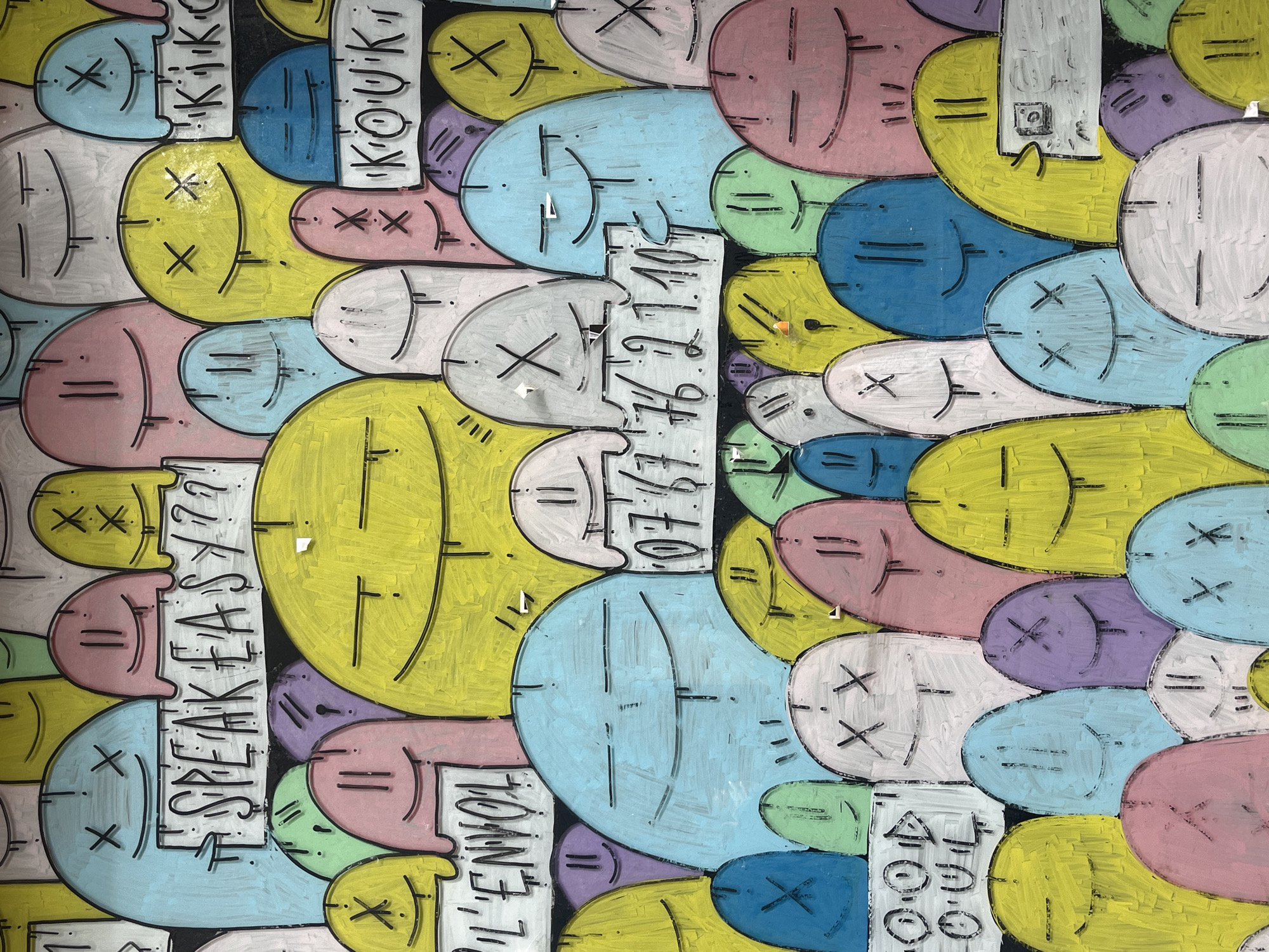 The image shows a close-up of a colorful, abstract mural or wall art. The artwork consists of many stylized faces painted in various pastel colors such as pink, blue, yellow, green, white, and purple. Each face is outlined with a black line, and they have simple facial features like "X"s for eyes, horizontal lines, or curved lines for mouths.
Interspersed among the faces are white rectangular blocks with black text written on them. The text includes phrases like "SPEAK EASY??", "L'ENVOL", "KOUK", "KIKIK", "HOANG., T., 0767 76.21.10-", "400", and "OUL". The text appears handwritten or stylized in a way that matches the overall aesthetic of the mural.
The image is a close-up, providing a detailed view of the textures and brushstrokes of the paint. Small pieces of tape or other objects are stuck to the painted surface in a few places.