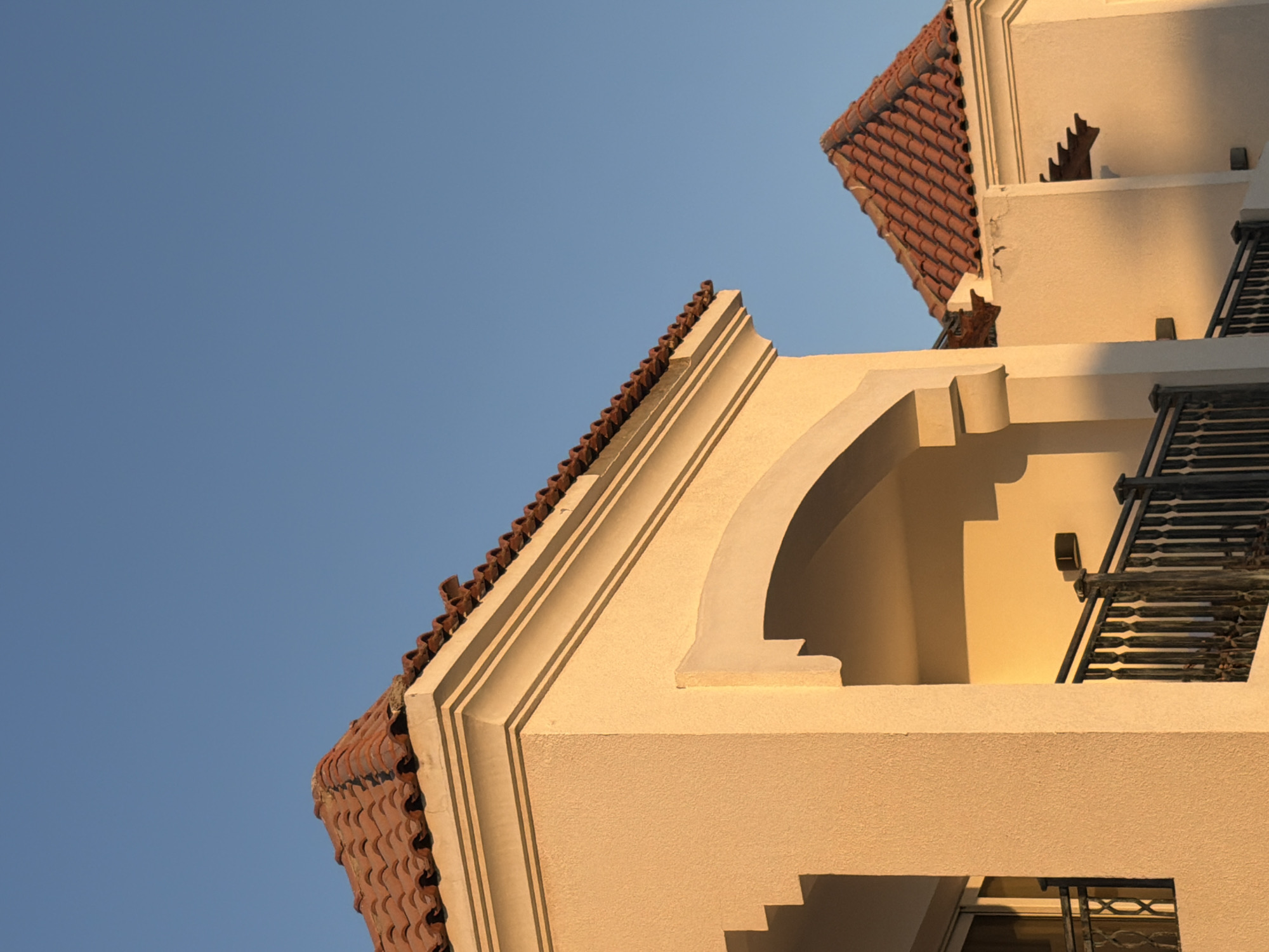 The image shows a close-up view of the terracotta tiled roofs and stucco architecture of a building against a clear blue sky. The sunlight casts warm shadows, suggesting it is either early morning or late afternoon. The architecture features ornate details, including decorative moldings and arched openings, hinting at a Mediterranean or North African style. A wrought-iron balcony railing is visible on the right side of the frame. The scene evokes a sense of tranquility and warmth, characteristic of a sunny, pleasant day. There is no visible text in the image.