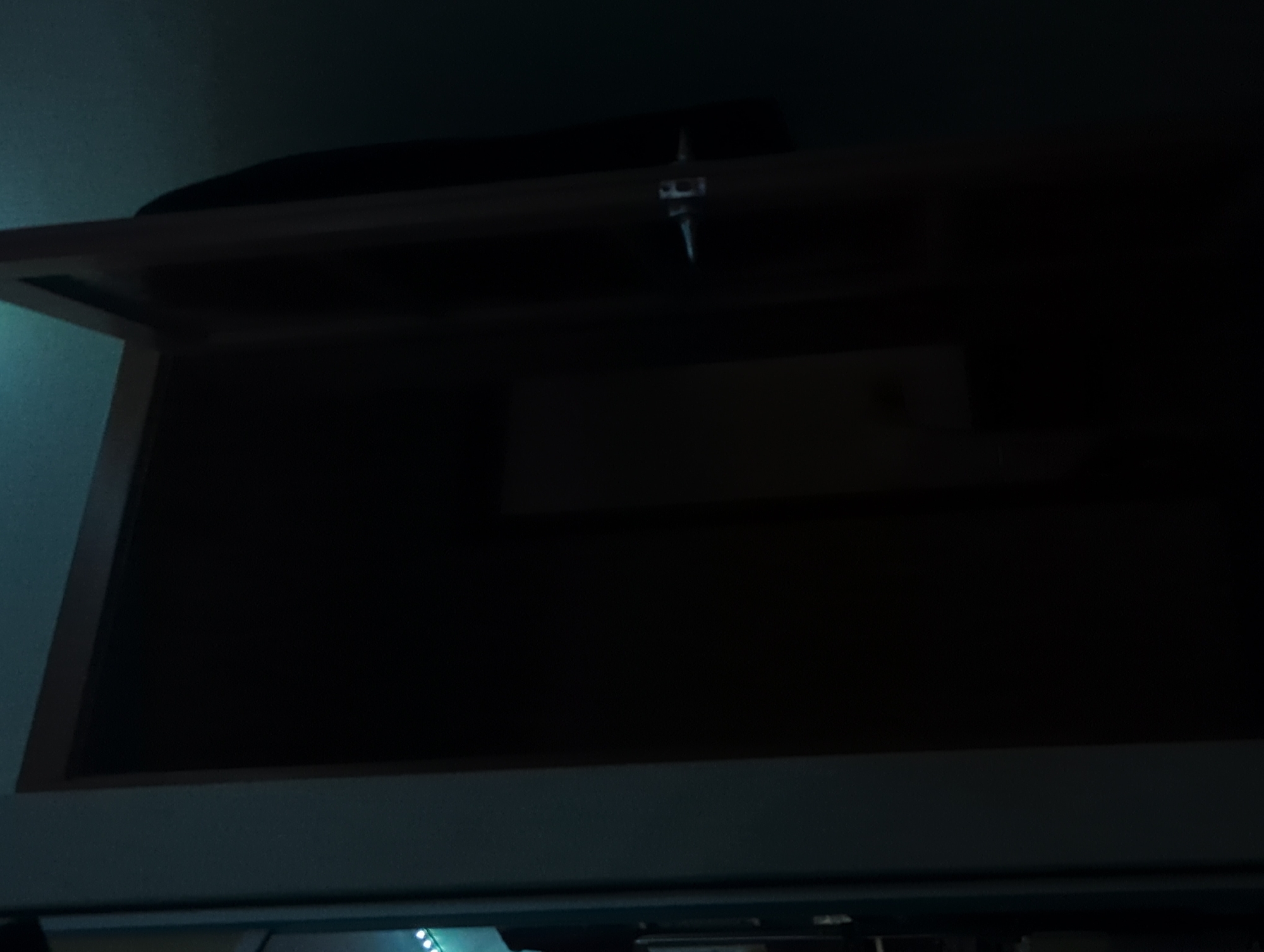 The image shows a slightly ajar dark wooden door with a silver doorknob. The door is partially lit by a faint teal light source from the left. The wall to the left of the door is light gray. A black object, possibly clothing, is hanging on the back of the door. The scene appears to be indoors, in a dimly lit environment. There is no visible indication of the city of Votkinsk, Russia, or any specific location within it. The image is a still photograph with no discernible action or interaction taking place.