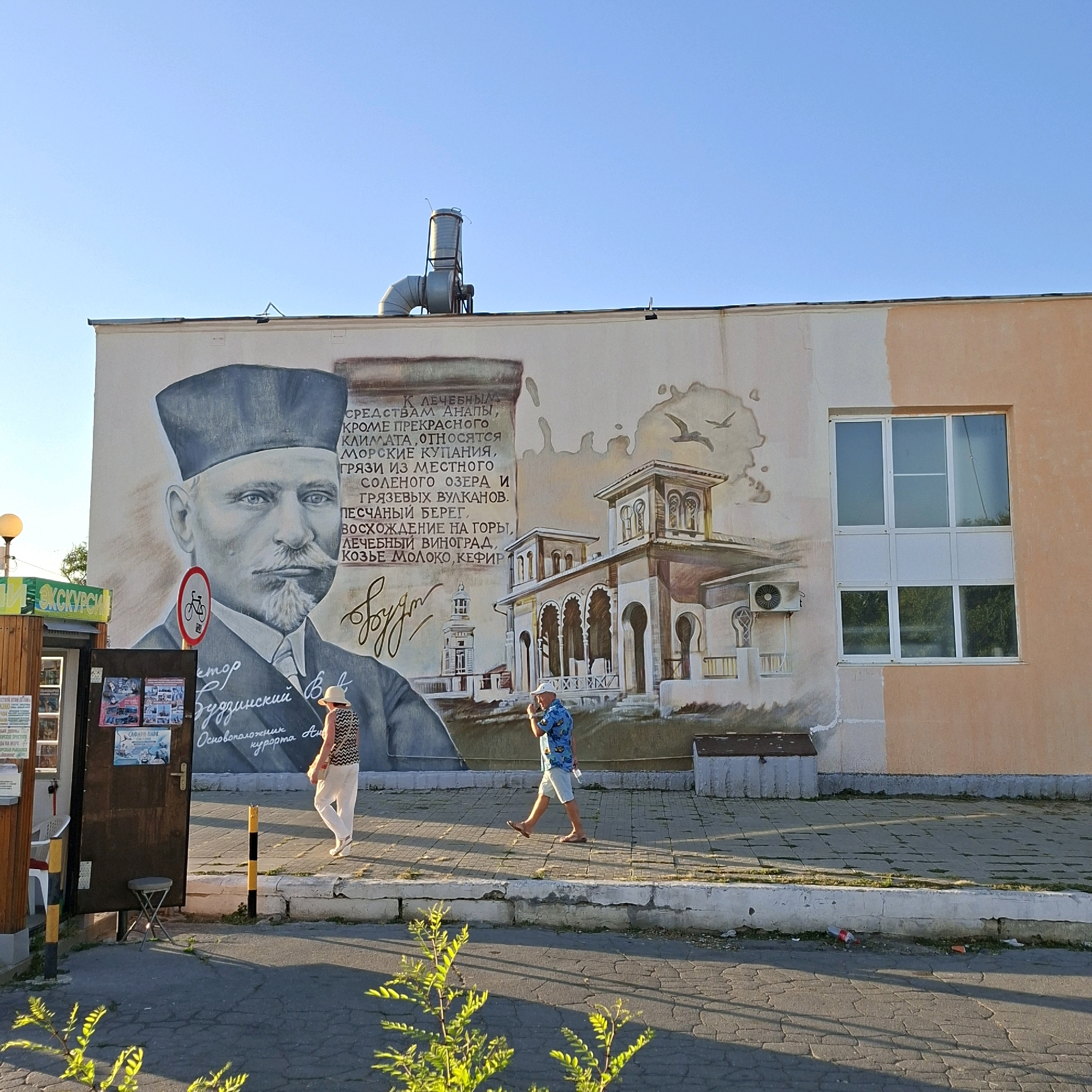 An outdoor, wide-angle, eye-level, full shot shows a mural on the side of a building. The mural depicts a portrait of a man in a hat and suit on the left, with text in Cyrillic next to him. To the right of the portrait is a drawing of a building with arched windows and a tower, and two birds flying above. A modern building with a large window is attached to the right side of the muraled wall. In front of the mural, two people are walking on a paved area. A woman in a patterned top and white pants walks away from the camera, while a man in a blue patterned shirt and shorts walks towards the camera. The ground is paved with grey squares, and a row of white concrete blocks runs along the base of the wall. There is a metal ventilation pipe on the roof of the building. Green plants are visible in the foreground and on the left side of the image. A street lamp is on the left.