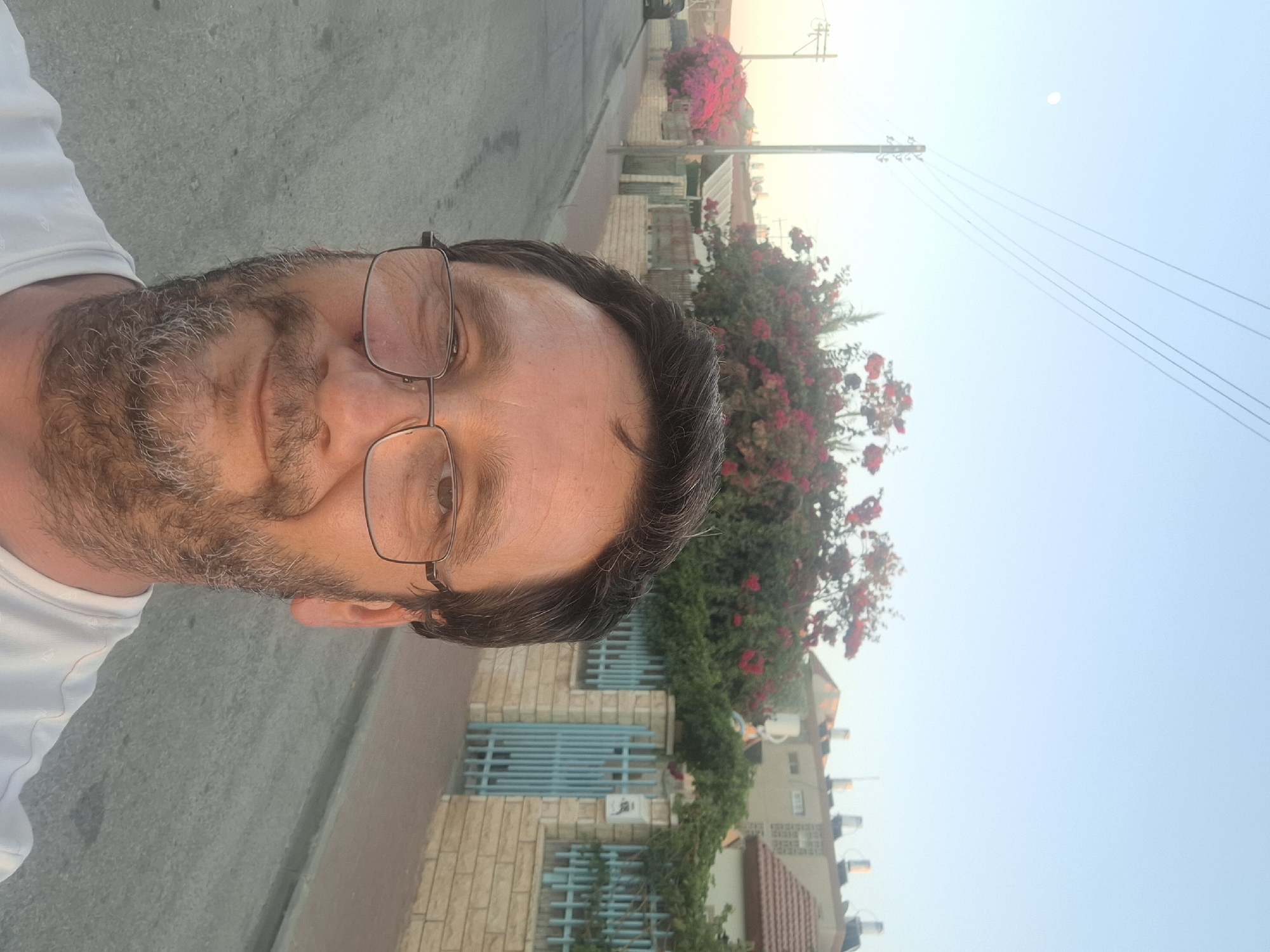 A medium-shot, selfie-style photograph captures a man with glasses and a salt-and-pepper beard looking directly at the camera with a slight smile. He is wearing a white t-shirt. The background depicts a residential street in Yerocham, Israel. A paved road runs parallel to the right side of the frame, with a sidewalk and a low brick wall bordering it. Beyond the wall, there is a pale blue metal gate and more brickwork. Lush green foliage with bright pink and red flowers adorns the area behind the wall. In the distance, residential buildings with tiled roofs are visible under a clear, pale blue sky. A telephone pole with wires and the moon are also present in the sky. The lighting suggests it is either early morning or late afternoon.