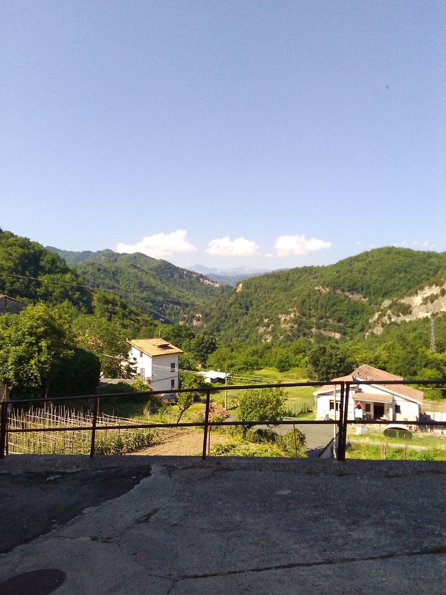 The image captures a scenic view of the mountainous landscape surrounding Valle Castellana, Italy. In the foreground, a concrete area transitions into a metal railing, behind which a small cultivated garden is visible. Further back, nestled among lush green trees, are two white houses with terracotta roofs. The houses appear to be residential dwellings. The background is dominated by rolling green hills and mountains, with a clear blue sky above, dotted with a few wispy clouds. The overall impression is one of rural tranquility and natural beauty. There are no people visible in the image.