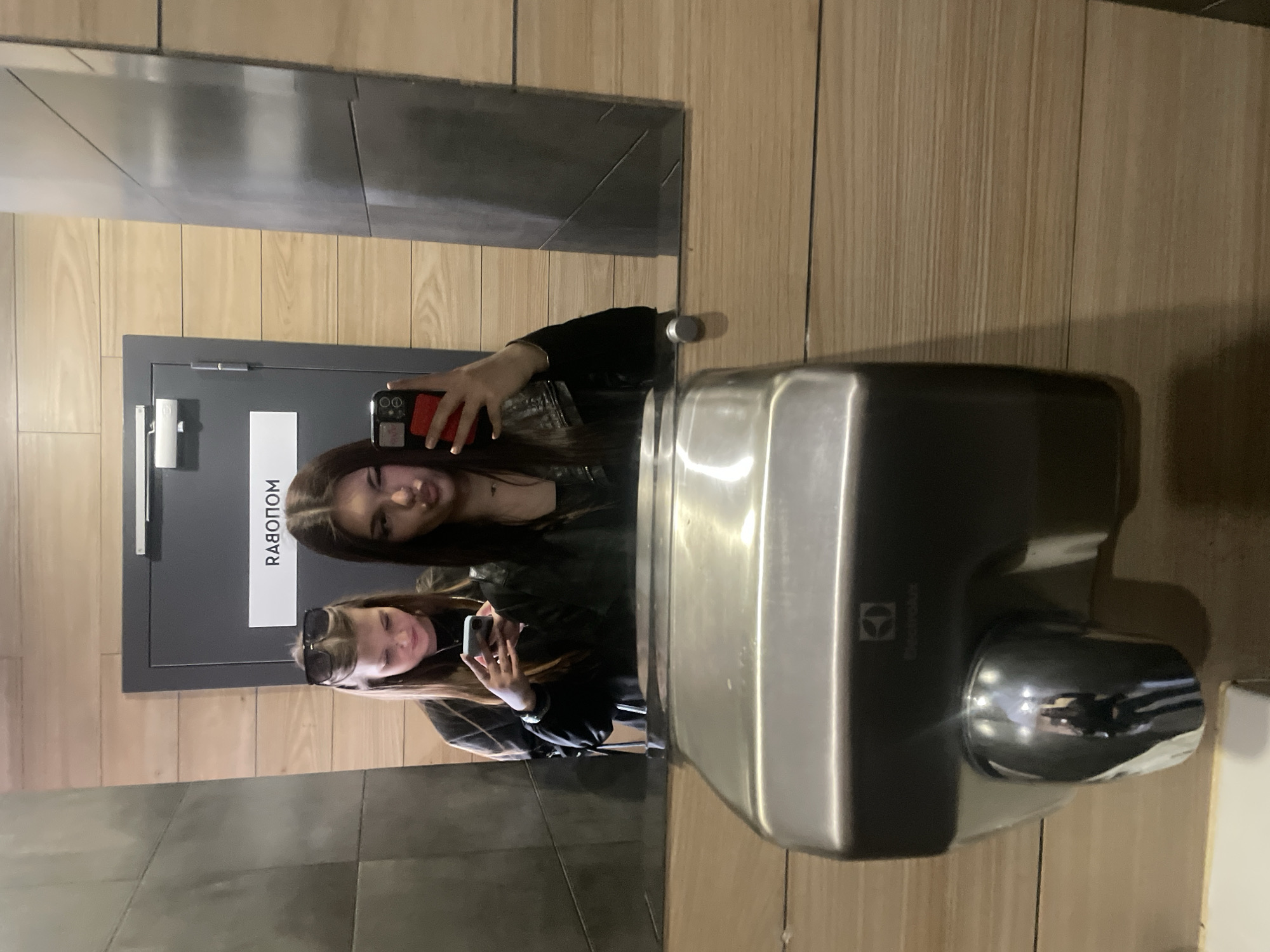 The image displays two young women reflected in a mirror or highly reflective surface, likely in a public restroom or changing room, as suggested by the presence of a hand dryer. The photo is rotated 90 degrees clockwise.
The main subjects are two women, both appearing to be taking a selfie. The upper woman (as seen in the rotated image) is holding a red-cased smartphone, extending her arm to capture the reflection. She has dark hair and is wearing a dark top, possibly a leather jacket. She is looking towards the camera with a subtle smile. The lower woman wears sunglasses on her head and is holding a white-cased smartphone in her hand, also wearing a dark top. She is also looking towards the camera with a slight smile. A smartwatch is visible on one of their wrists.
The setting features light wooden paneling on the walls and dark gray or black rectangular tiles on another surface, possibly the floor or a counter. A dark gray door is prominent in the background, featuring a light-colored handle. On the door, there is a white rectangular sign with mirrored Russian text, which appears to read "RABOTOM" (РАБОТОМ). This is likely a reflection of "РАБОТАЕТ" (Rabotaet), meaning "is working" or "open."
To the right of the women, a metallic, modern hand dryer is mounted on the wall. The Electrolux logo and brand name are visible on its side. The hand dryer has a reflective nozzle, showing faint reflections of the surrounding area. The overall lighting suggests an indoor setting with artificial light. The activity is a casual selfie-taking moment between the two individuals.
