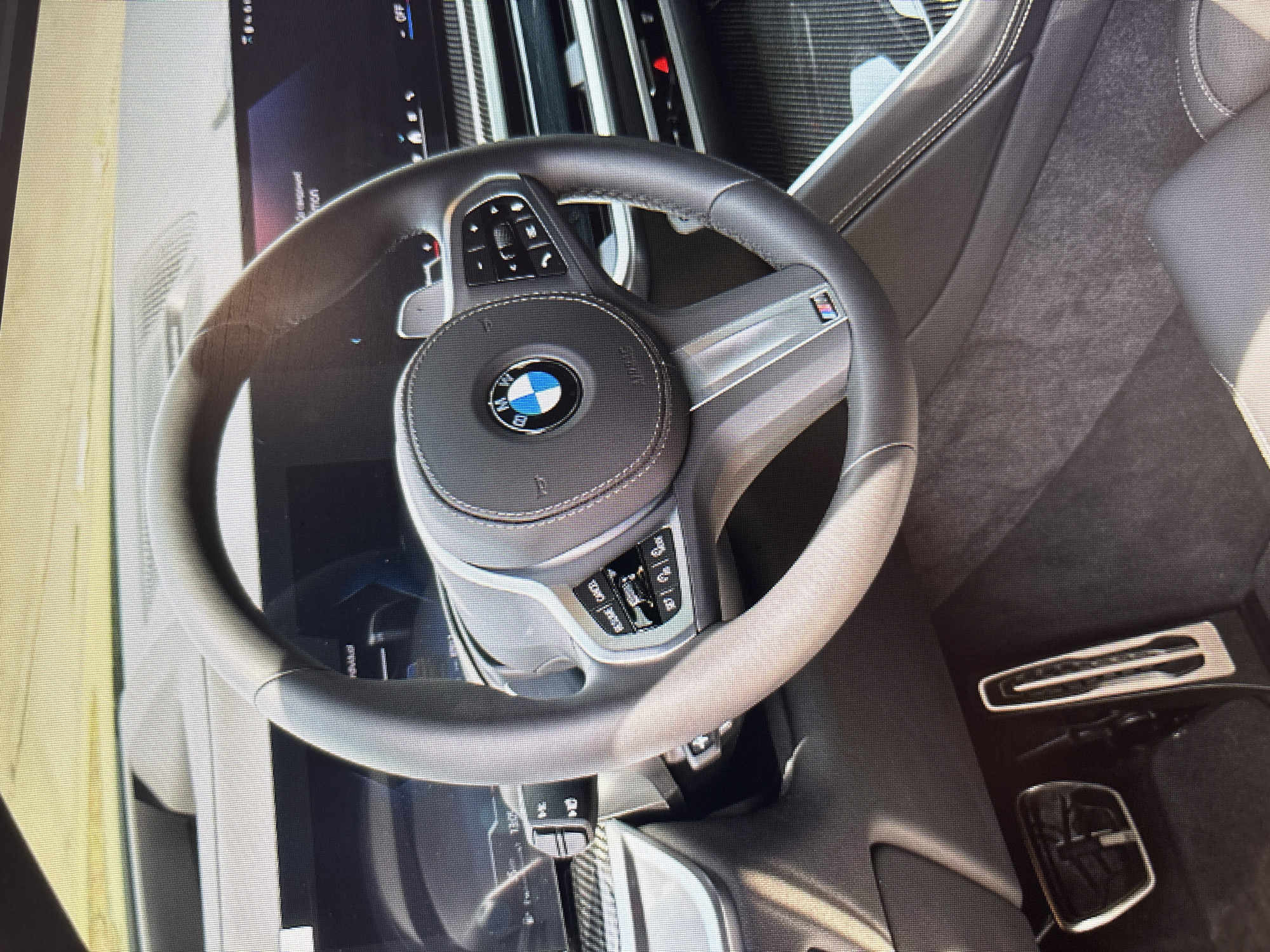 The image shows a close-up, interior view of a modern car, specifically the driver's side. The steering wheel, featuring the BMW logo and M-sport badging, is prominently displayed. Various controls are visible on the steering wheel, including buttons for cruise control and audio. To the left of the steering wheel, a turn signal lever and a digital instrument cluster displaying some information can be seen. Below the steering wheel, the accelerator and brake pedals are visible. The dashboard and center console are also partially visible, with a carbon fiber trim accent. The overall aesthetic suggests a luxury vehicle. There is no indication of the car's location in Moscow, Russia, or any other specific geographical area. The image is static and does not depict any ongoing actions or interactions.