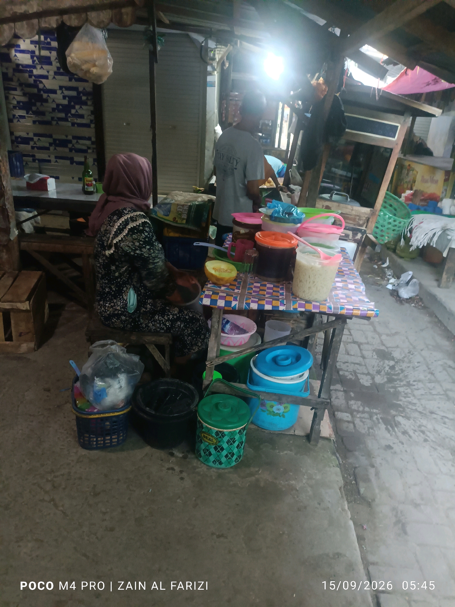 The image captures a street-side stall in Arosbaya, Indonesia, likely a food or drink vendor. A woman wearing a pink hijab and a patterned dress is seated on a stool behind a table laden with various containers. She is facing away from the camera, and her left hand appears to be interacting with something on the table. To her right, a portion of a light-colored melon is visible on a colorful patterned cloth.
Several containers are present on the table, including large orange and dark-colored tubs with lids, a clear plastic cup filled with a light-colored, textured liquid and a pink spoon, and several smaller bowls. A green bucket with a lid and a diamond pattern is positioned on the ground in front of the table. To the left of the woman, a blue basket filled with items, including plastic bags and containers, is visible. A black bucket is also on the ground nearby.
In the background, a person wearing a grey t-shirt stands behind the table, facing left. Further in the background, the scene shows stall structures, closed metal shutters, and hanging items, suggesting a market or a row of shops. The ground is paved and appears wet in places. Overhead, wooden beams and corrugated roofing are visible, with some pink fabric draped over parts of the structure. A green bottle is also present on a surface behind the woman. The overall lighting suggests it is either early morning or late evening, with a strong light source emanating from the upper left. The timestamp "15/09/2026 05:45" is overlaid on the bottom right corner of the image.
