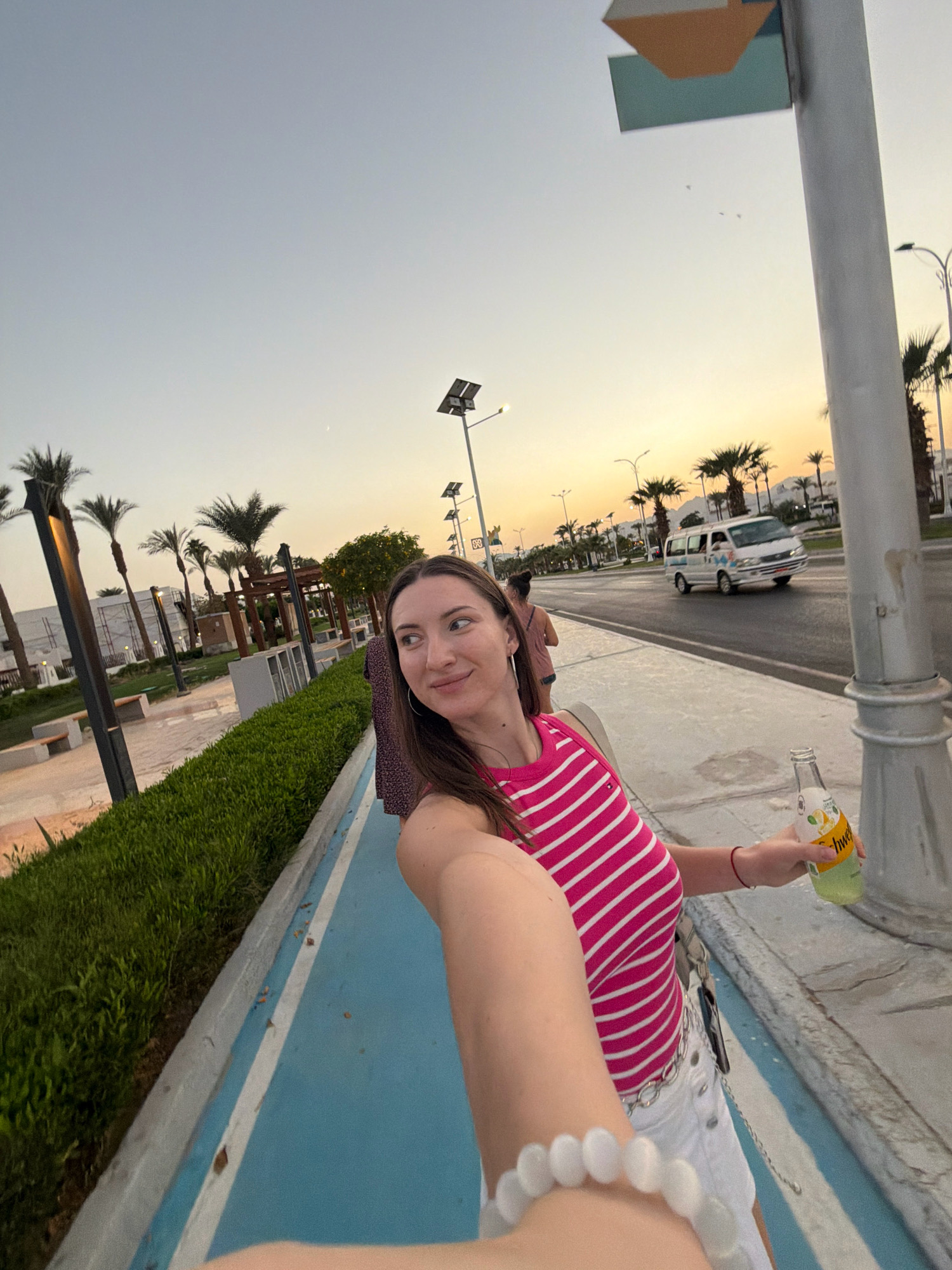A woman, wearing a pink and white striped tank top and white shorts, takes a selfie while holding a bottle of Schweppes. She is standing on a sidewalk next to a blue path and a row of green bushes. In the background, palm trees and buildings line a street where a white van is driving. The sky is a pale orange, indicating either sunrise or sunset, and a faint crescent moon is visible. The location is the city of Sharm El Sheikh, Egypt.