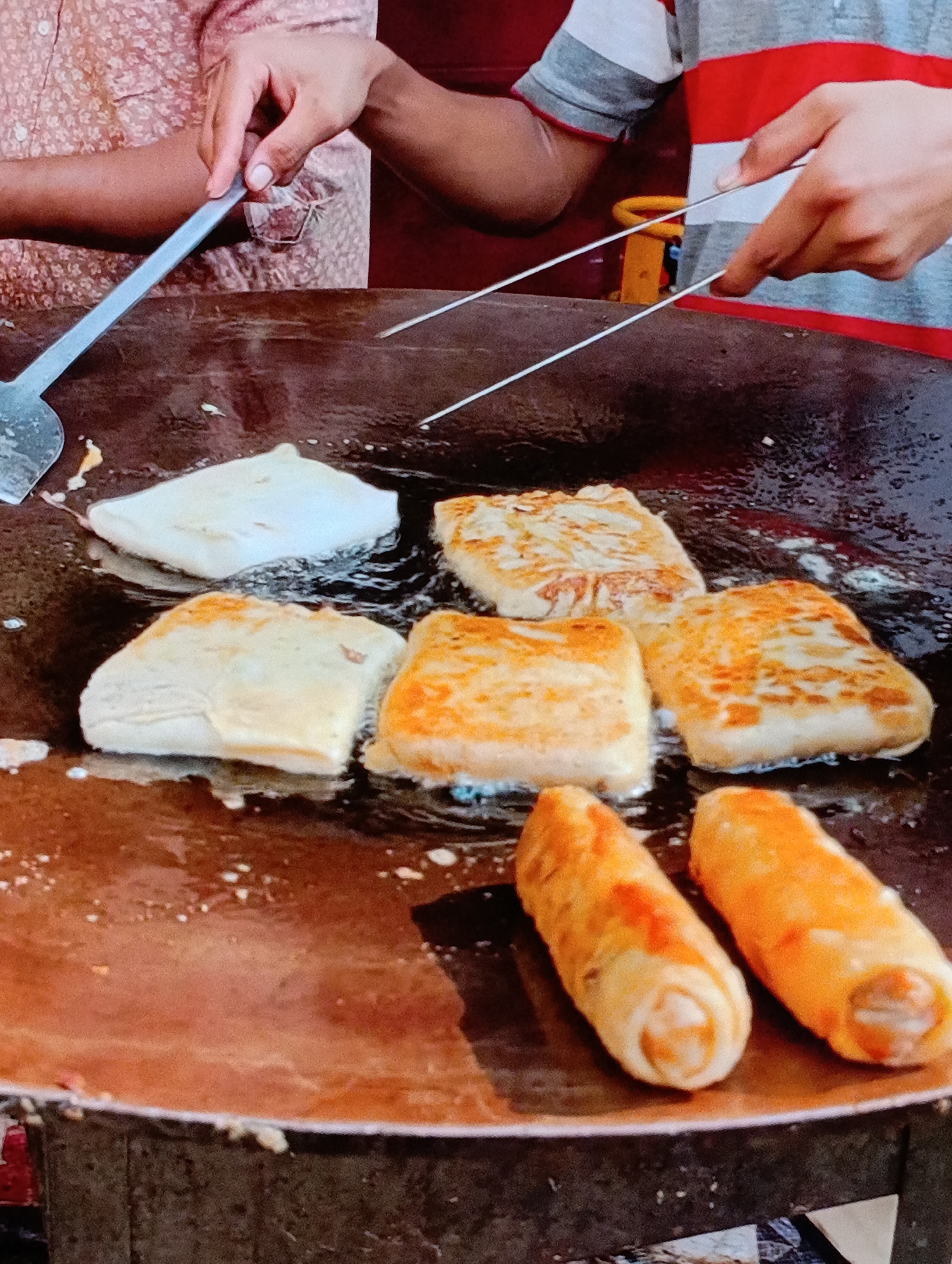 Captured at an unknown location in the city of Unknown, the image presents a scene of food preparation. A large, circular, dark-colored flat griddle or pan is the primary focus, with several items being cooked. At the top left, a hand holds a spatula, while another hand on the top right is maneuvering thin, metal tongs. Five square-shaped foods with varying degrees of browning are arranged across the pan, seemingly fried. In the foreground, two cylindrical objects, appearing to be rolled food items, are positioned. The overall environment suggests a street food setting.